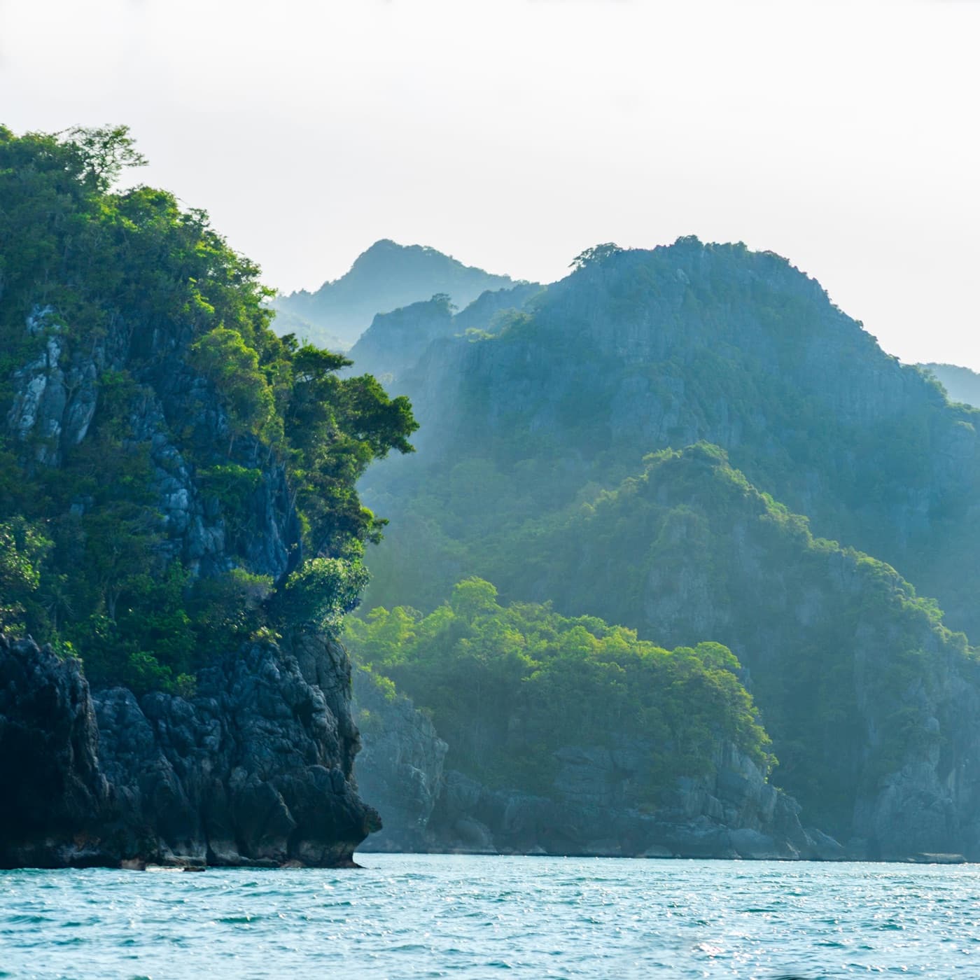 Steep, forested limestone islands rising from a choppy blue-green sea in Ang Thong Marine National Park, the farther peaks fading into a pale haze