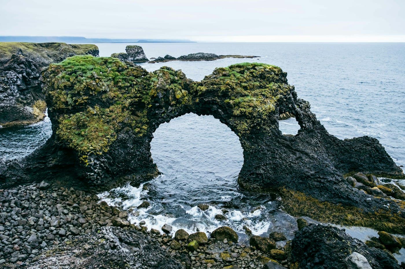 Gatklettur, un arco de basalto cubierto de musgo en Arnarstapi, con el mar entrando por debajo hasta una orilla de cantos rodados
