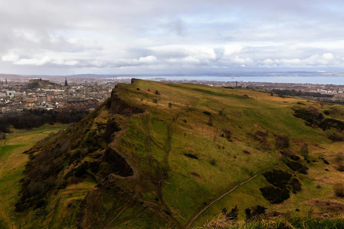 La cresta verde de Salisbury Crags vista desde Arthur’s Seat, con Edimburgo, el castillo y el estuario del Forth al fondo
