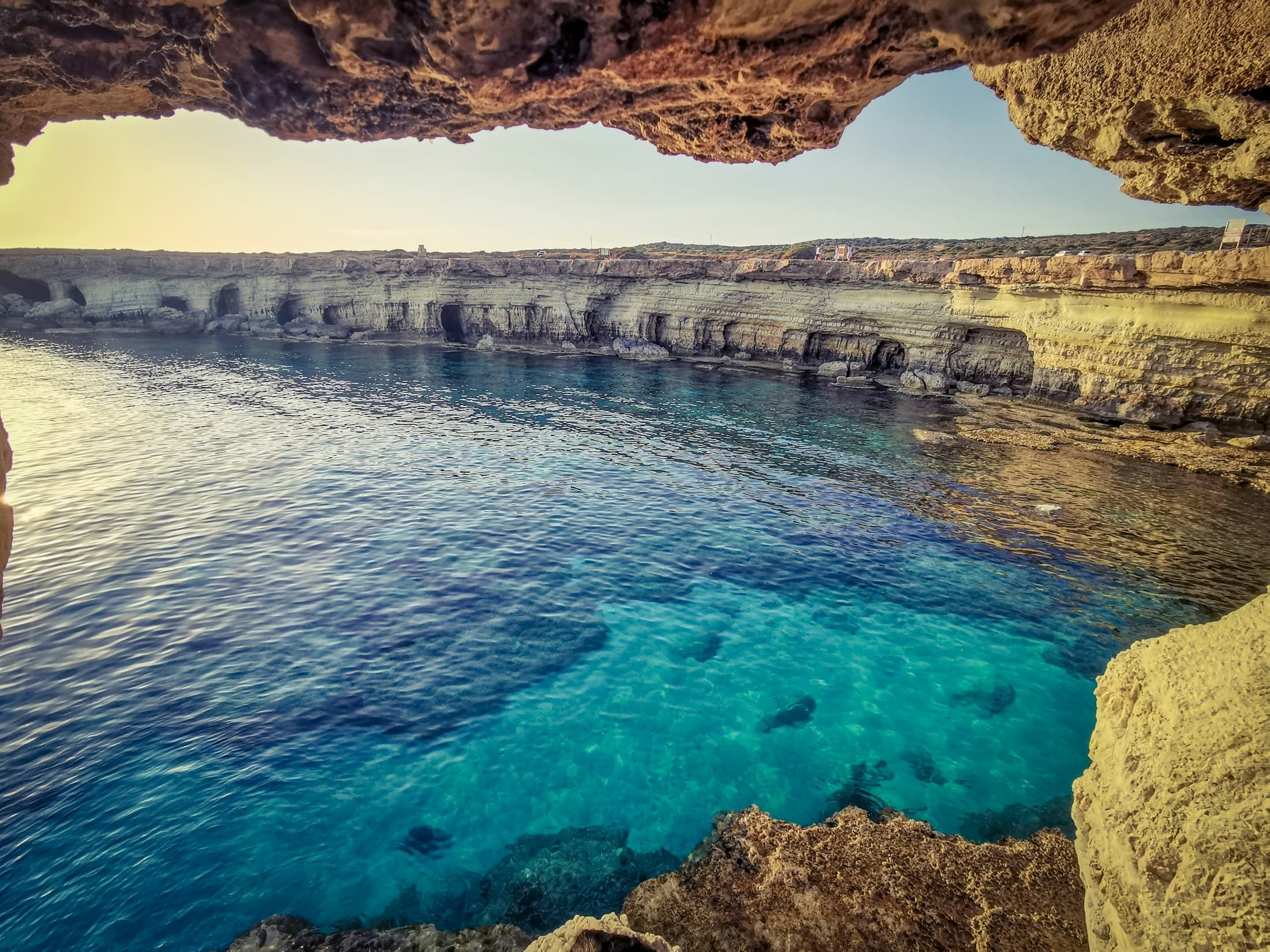 The sea caves near Ayia Napa framed by the roof of a cave: turquoise water and a long line of pale cliffs hollowed with caves, in warm late light