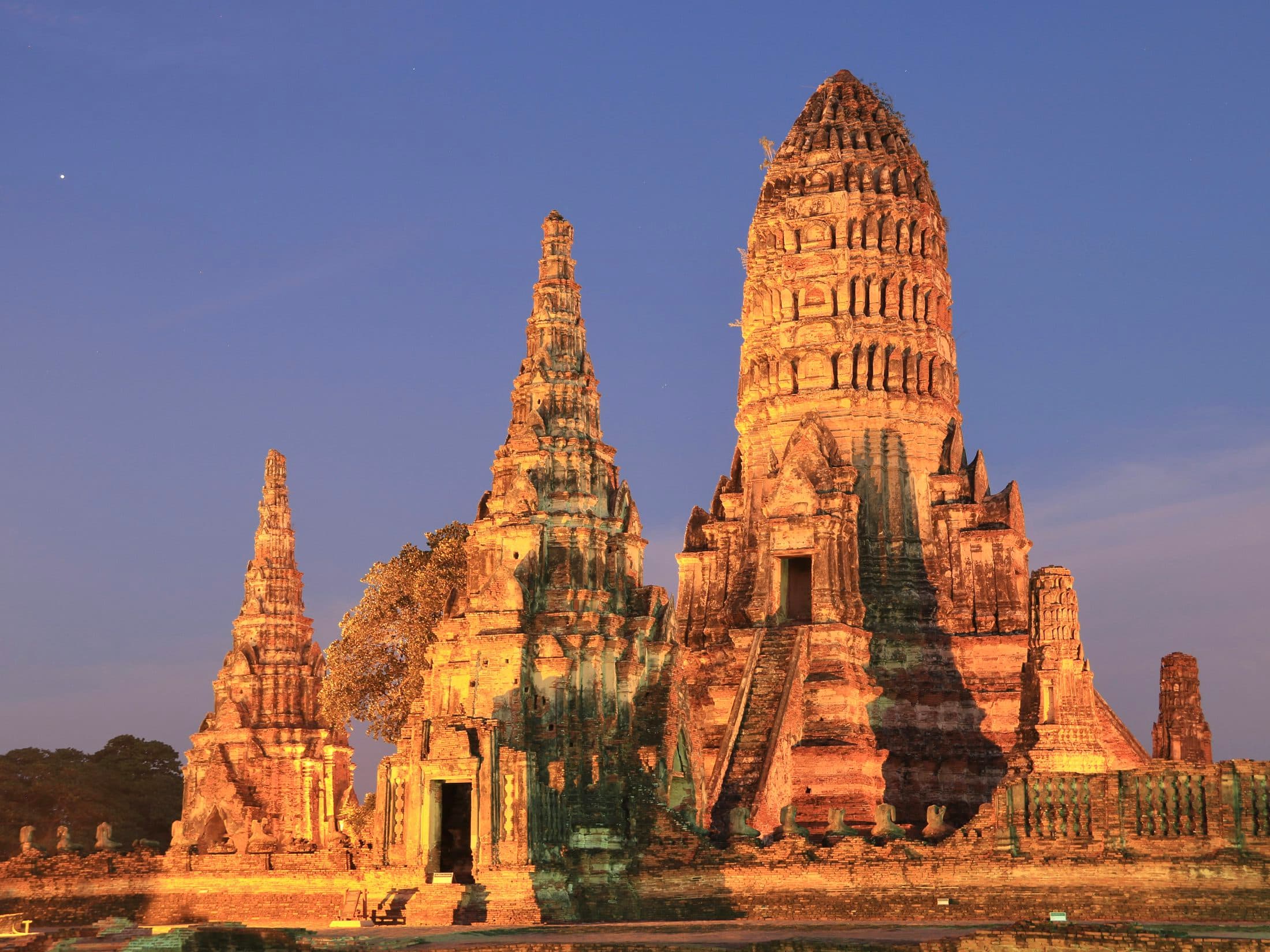 Die Backstein-Prangs des Wat Chaiwatthanaram in Ayutthaya, in der Abenddämmerung warm golden angestrahlt, der hohe Mittelturm mit seiner steilen Treppe zwischen kleineren Türmen unter einem klaren blauen Abendhimmel