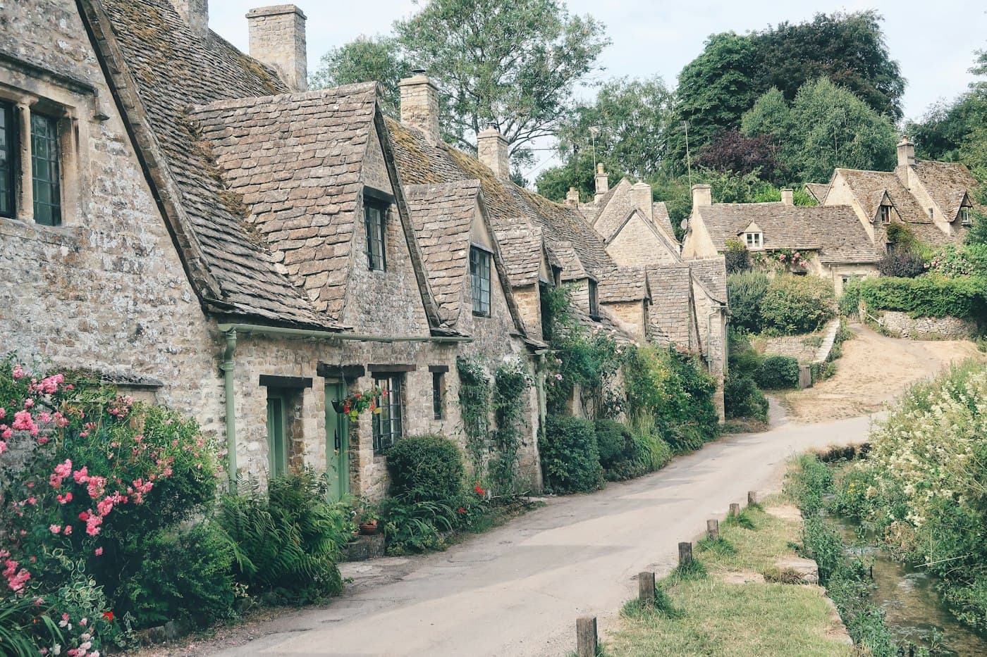 Arlington Row in Bibury, stone weavers’ cottages with mossy stone-tiled roofs and climbing roses along a narrow lane, pink roses in front, a little stream and the grassy edge of Rack Isle on the right and more cottages up the hill