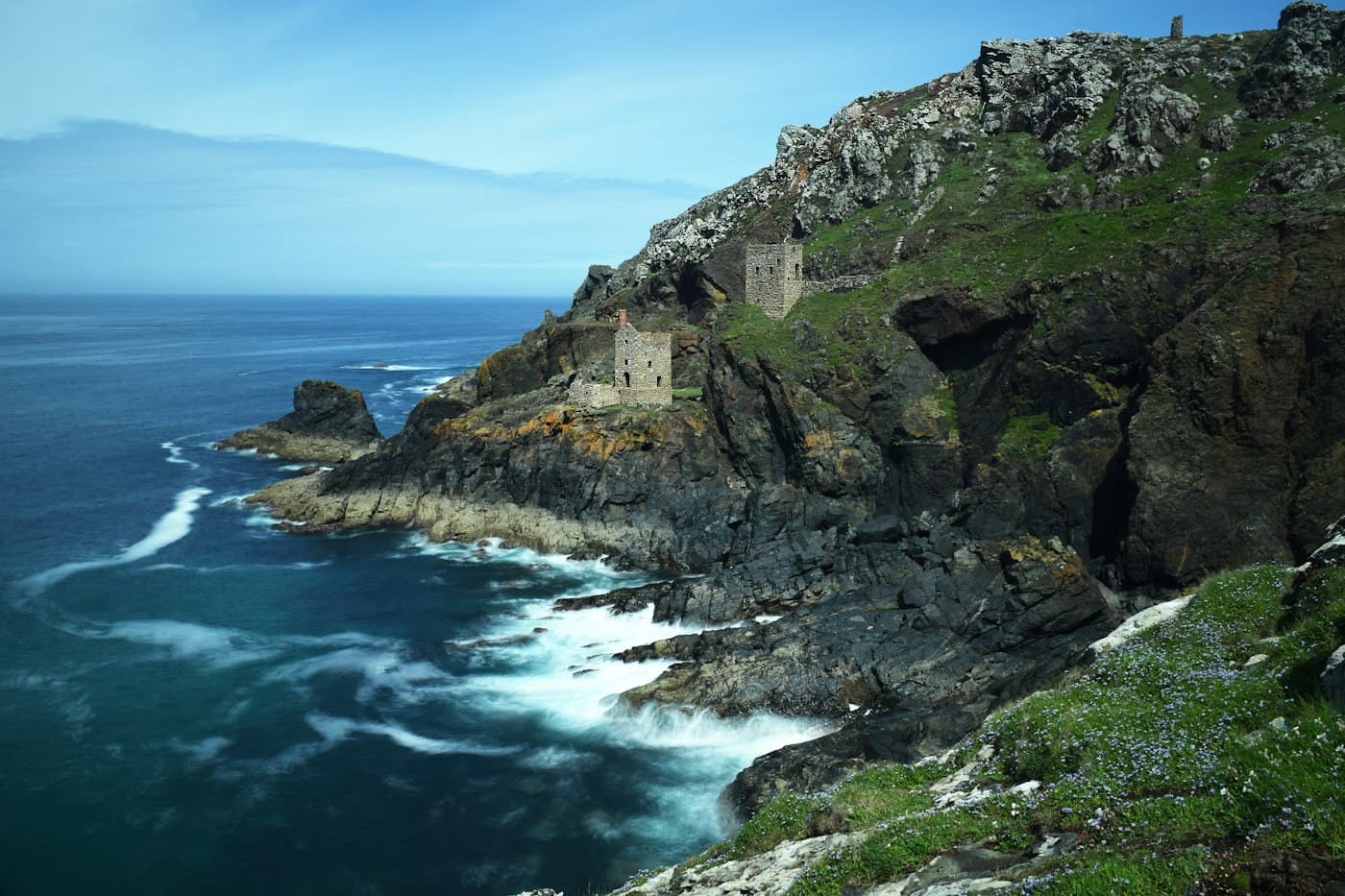 I due edifici in rovina della miniera Crowns, uno sopra l’altro sulle scogliere scure di Botallack, visti dal sentiero costiero, sopra un mare blu intenso striato di schiuma bianca, con piccoli fiori azzurri nell’erba in primo piano