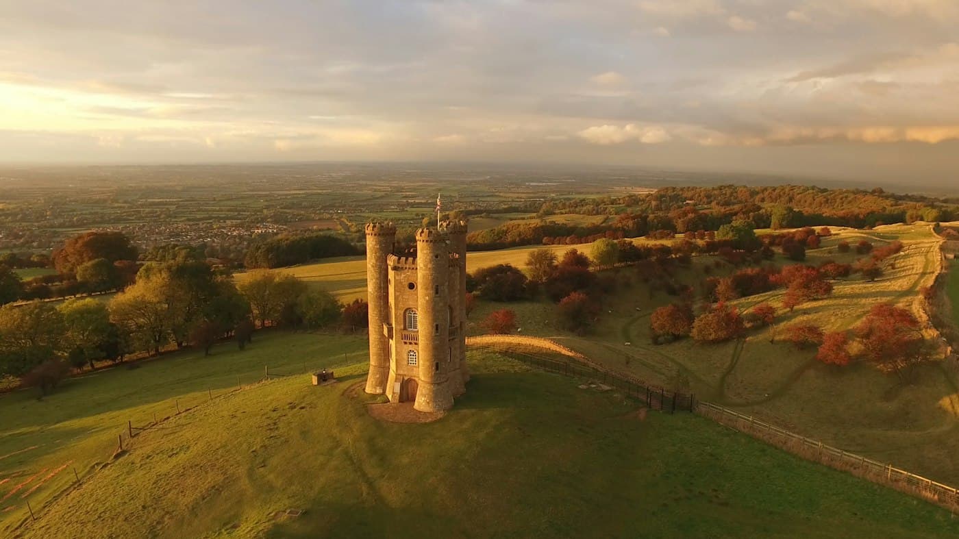 Broadway Tower, a turreted stone folly on the grassy top of Beacon Hill, seen from above and lit gold by low autumn sun, with copper-leaved trees on the slopes and the plain stretching to the horizon