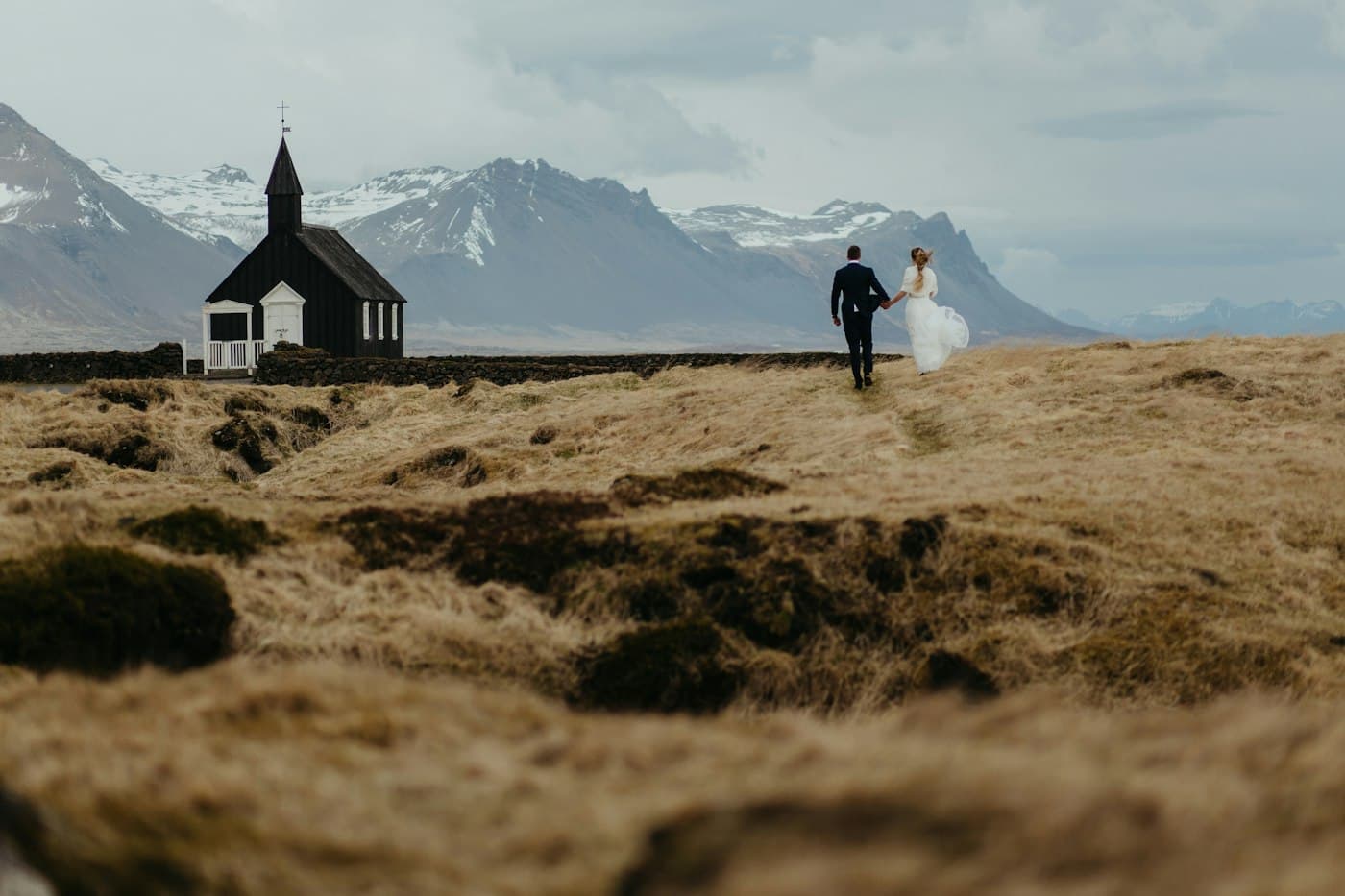 Unos novios cogidos de la mano cruzan un campo de lava cubierto de musgo hacia la iglesia negra de Búðir, con montañas surcadas de nieve detrás