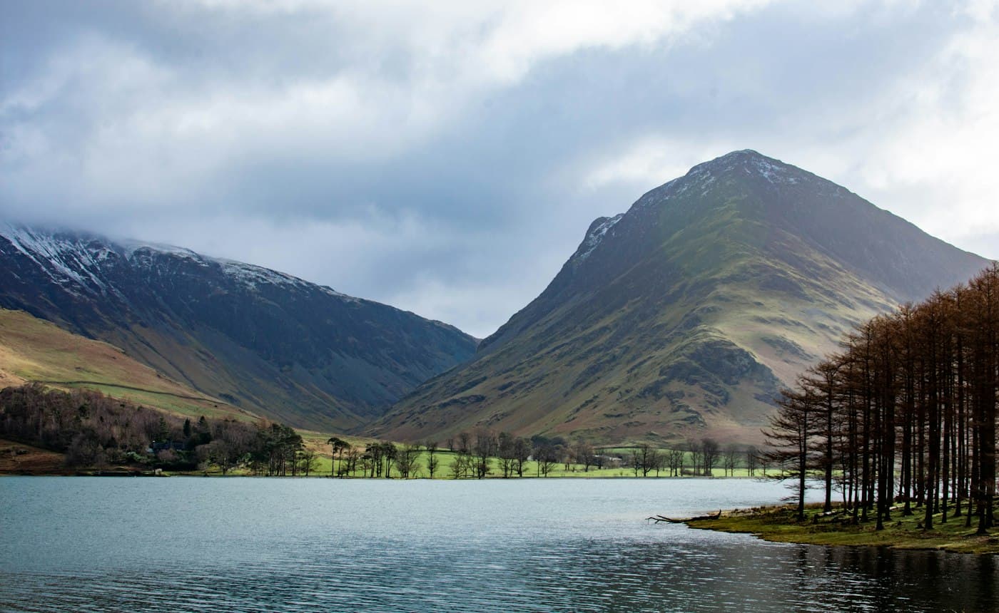 Buttermere under heavy cloud, the pointed peak of Fleetwith Pike rising beyond the lake, a stand of tall bare trees on the shore and snow on the higher fells