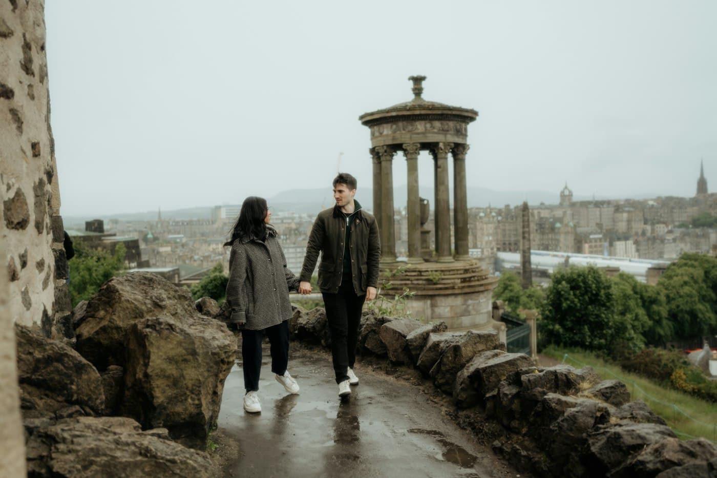 Una pareja de la mano por un camino mojado de Calton Hill, junto al templete redondo de columnas del Dugald Stewart Monument, con los tejados y las agujas de la Ciudad Vieja detrás