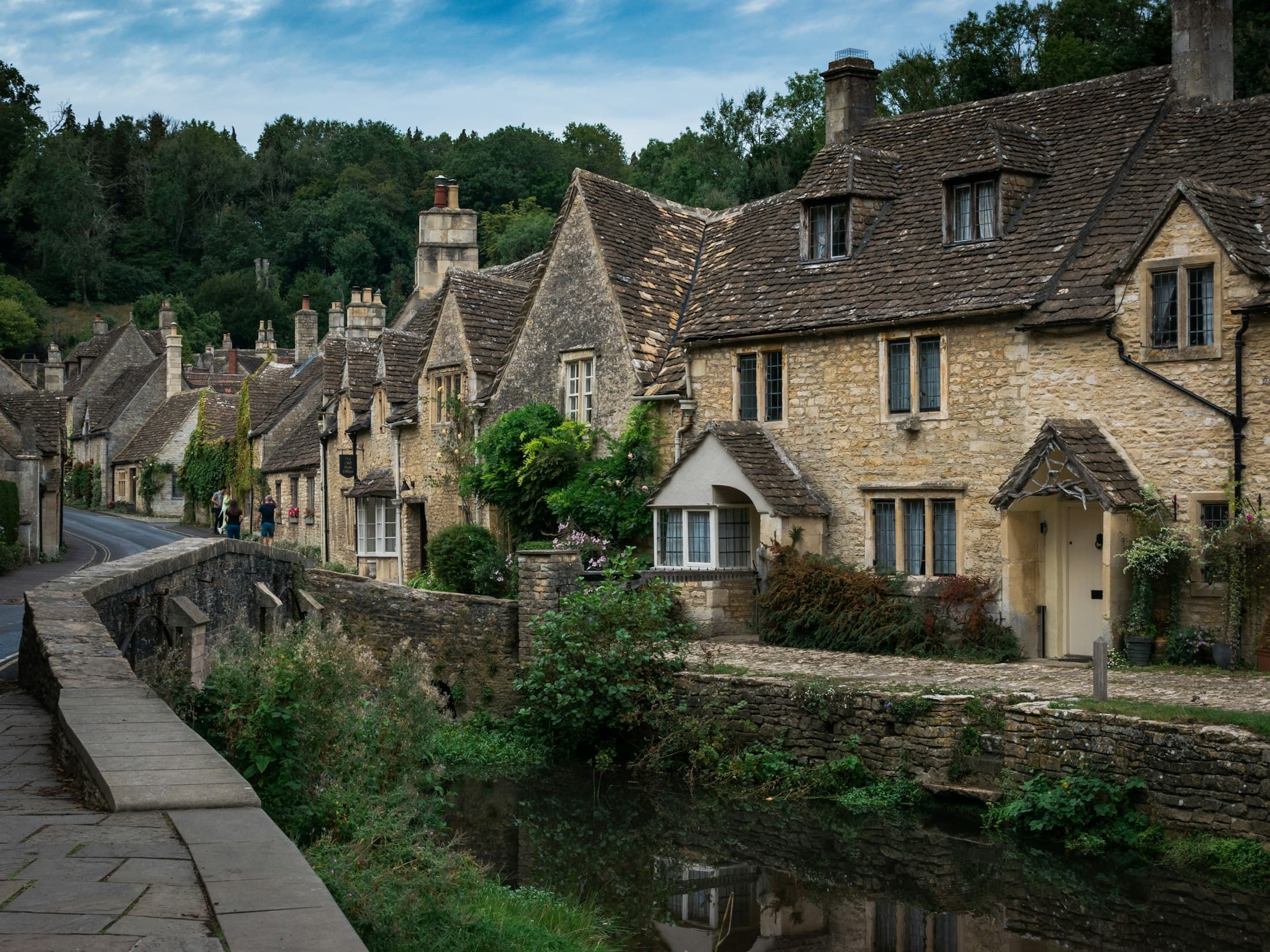 Steinhäuser mit steilen Dächern aus Steinplatten und Gauben am By Brook in Castle Combe, links die Brüstung der alten Brücke, dahinter die Dorfstraße, die zwischen weiteren Häusern zu bewaldeten Hängen ansteigt, unter blauem Himmel mit leichten Wolken