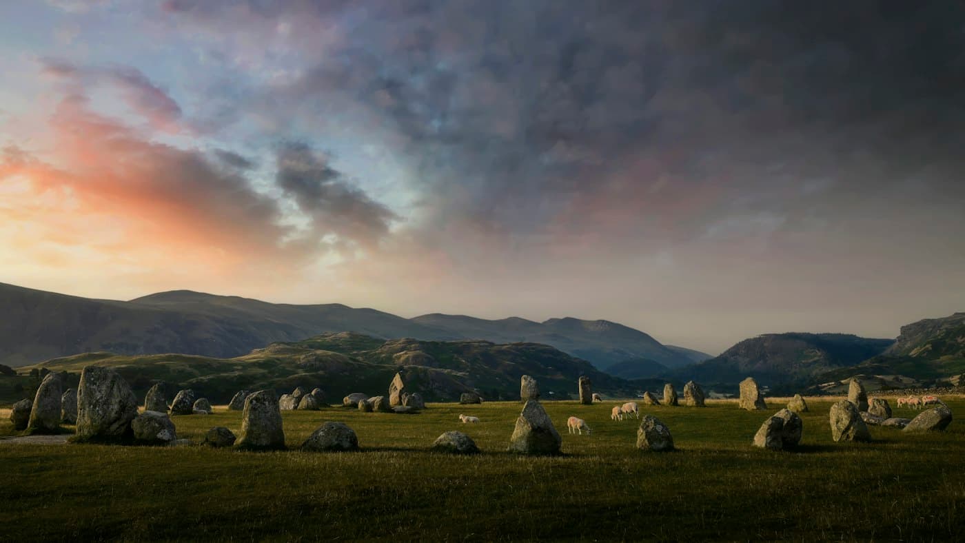 The standing stones of Castlerigg in low sunlight, sheep grazing among them and the fells beyond under heavy cloud tinged with pink