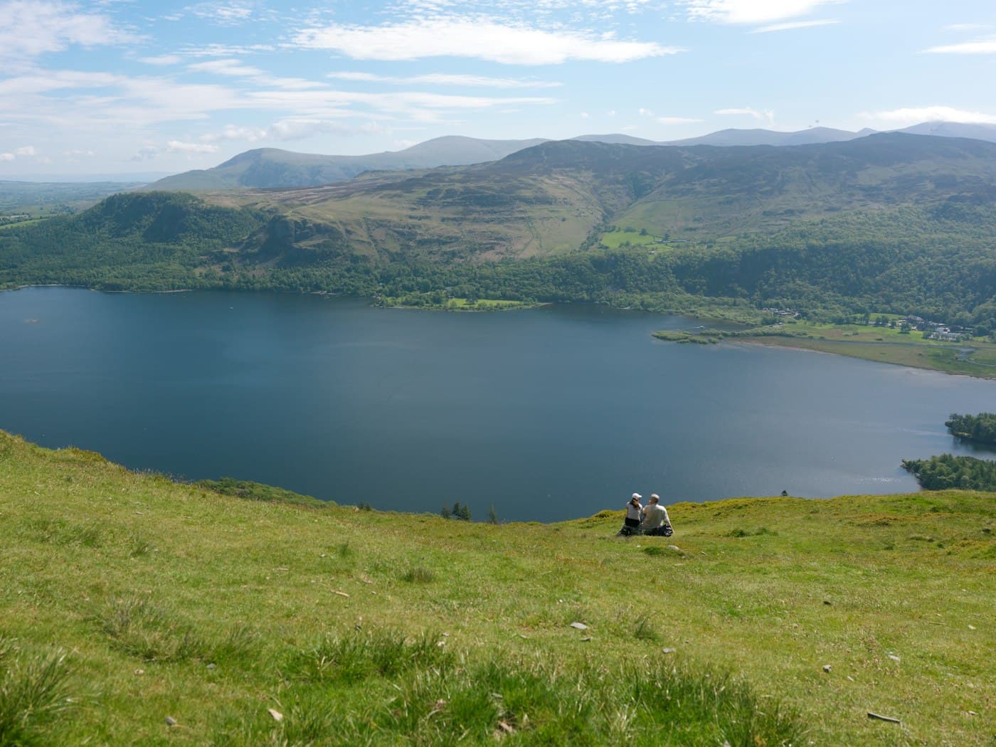 A couple sitting together on the grassy slope of Catbells, looking out over Derwentwater to the wooded shore and the fells