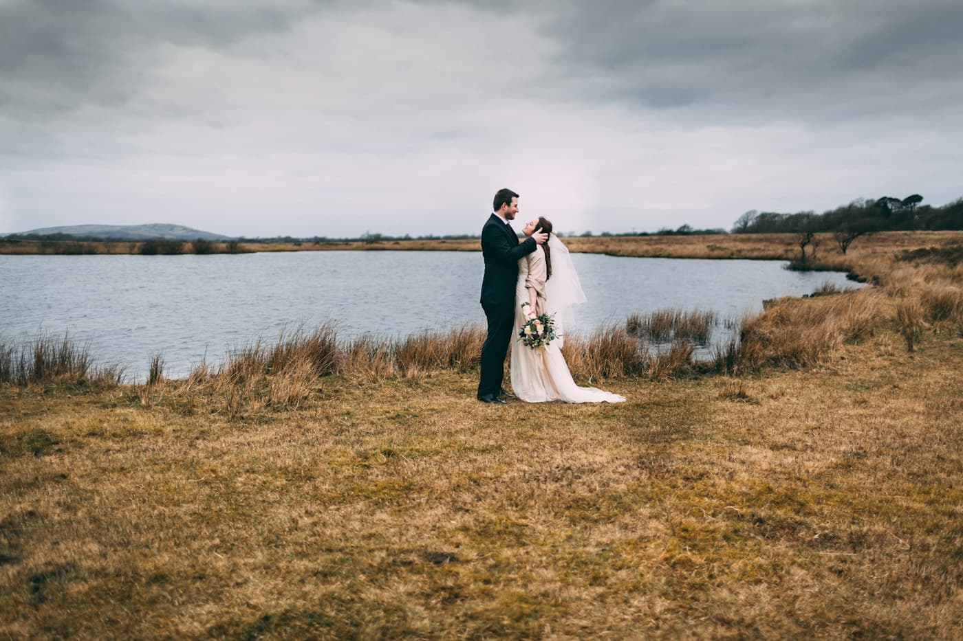 A bride and groom embracing by a moorland pool on Cefn Bryn, Gower, under a grey sky