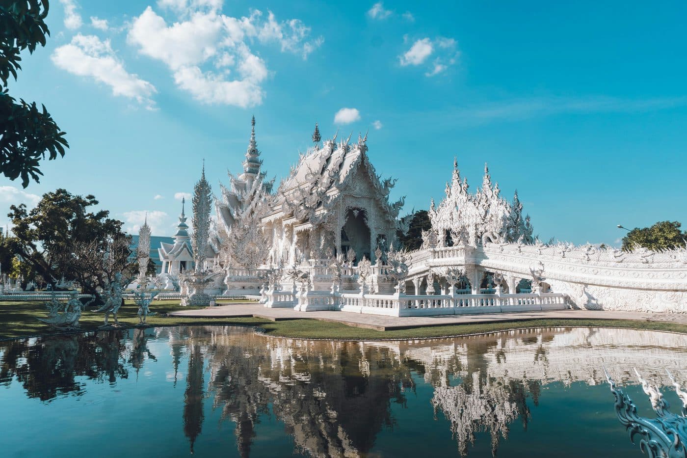 Die weißen Hallen und Spitzen des Wat Rong Khun, des Weißen Tempels von Chiang Rai, gespiegelt im stillen Wasser seines Teichs unter blauem Himmel mit weißen Wolken