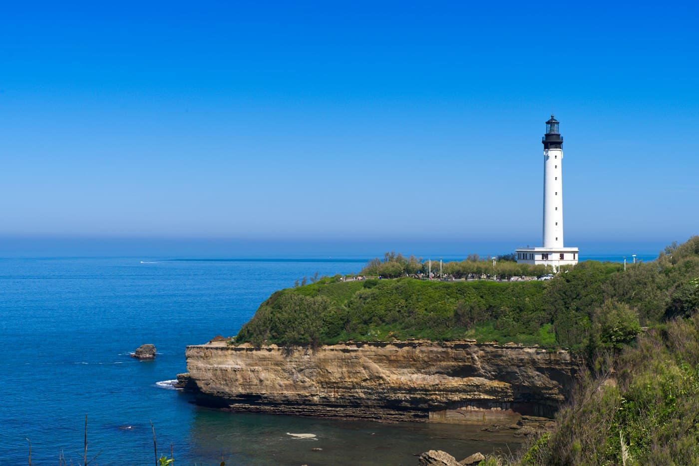 The white lighthouse of Biarritz on the green Pointe Saint-Martin, golden rock cliffs dropping into a deep blue sea