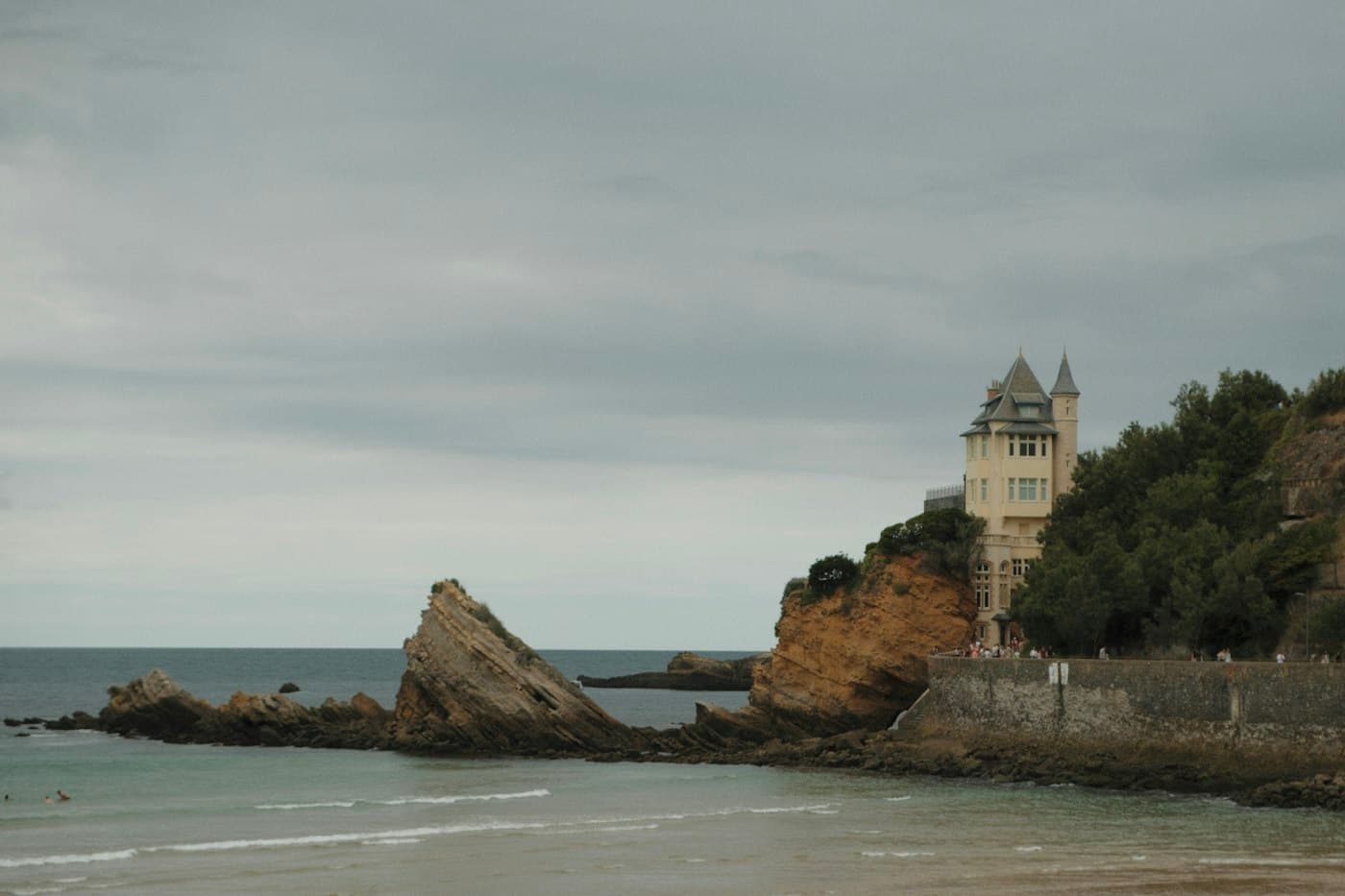 A turreted villa on a rocky point above the beach at Biarritz, jagged rocks running out into a grey-green sea under a cloudy sky