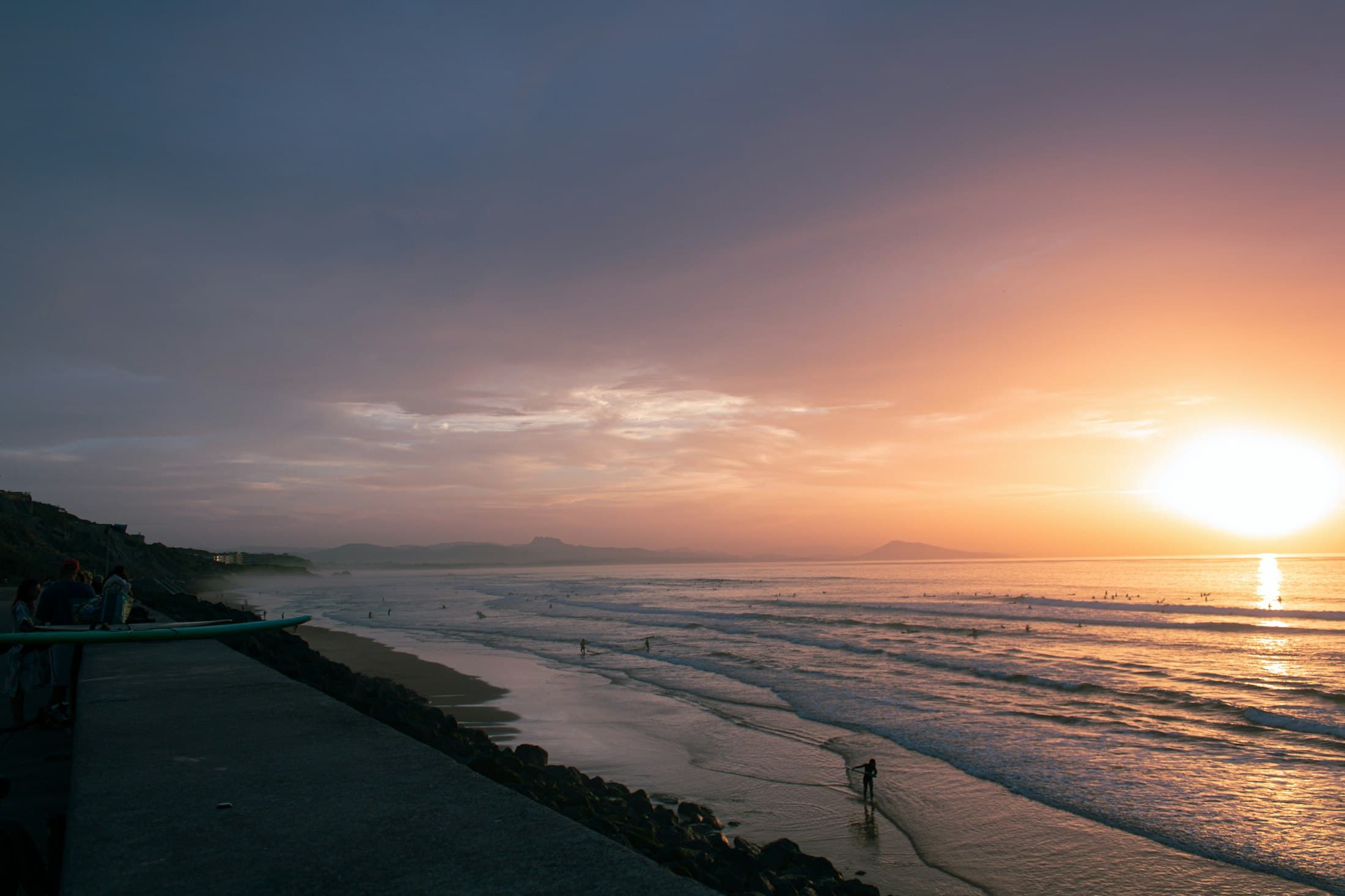 Sunset over the Atlantic at Biarritz, surfers in the low waves, a long beach under the cliffs and the Basque mountains on the horizon