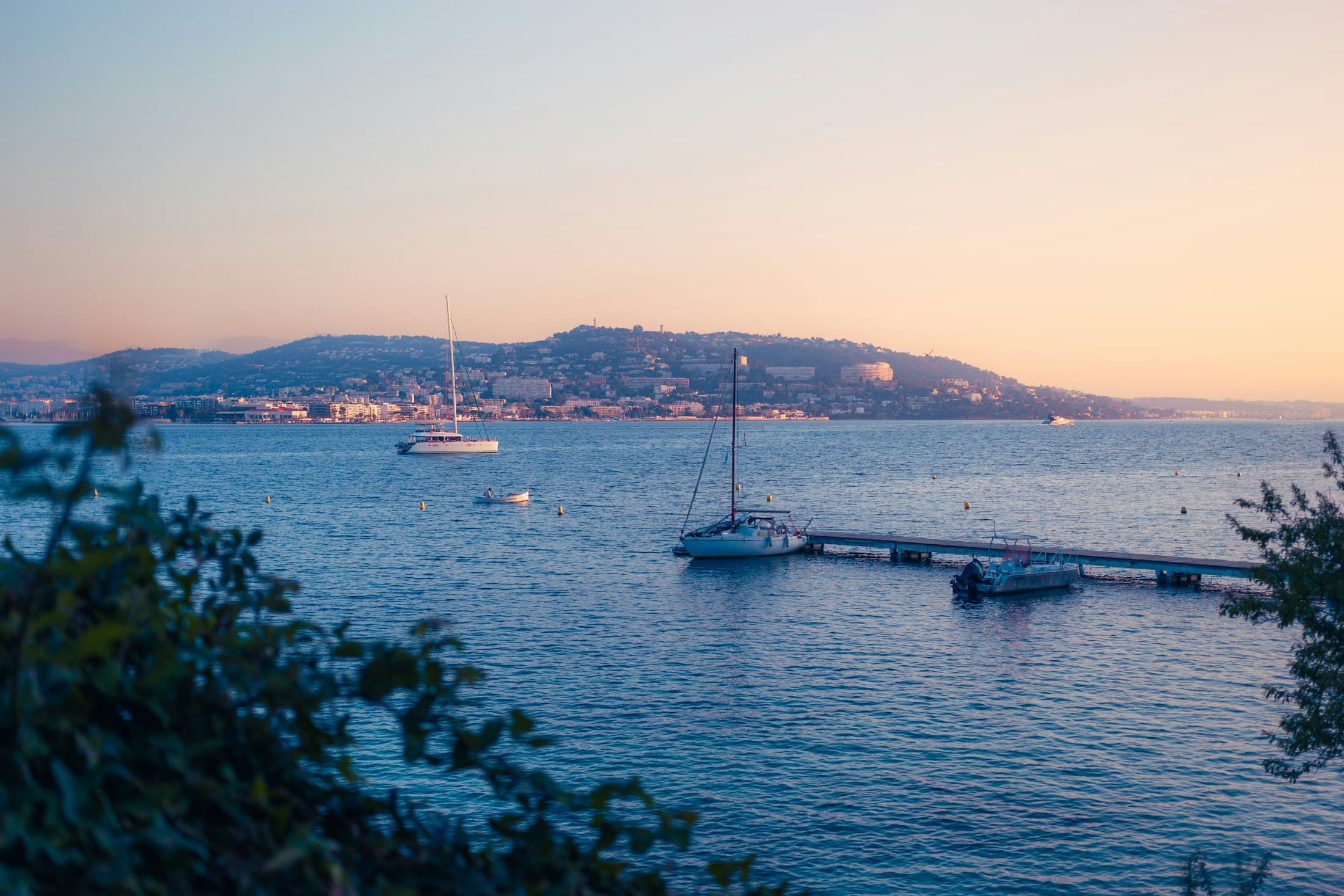 Cannes across the bay at dusk from the island of Sainte-Marguerite, sailing boats at anchor by a small jetty under a peach-coloured sky