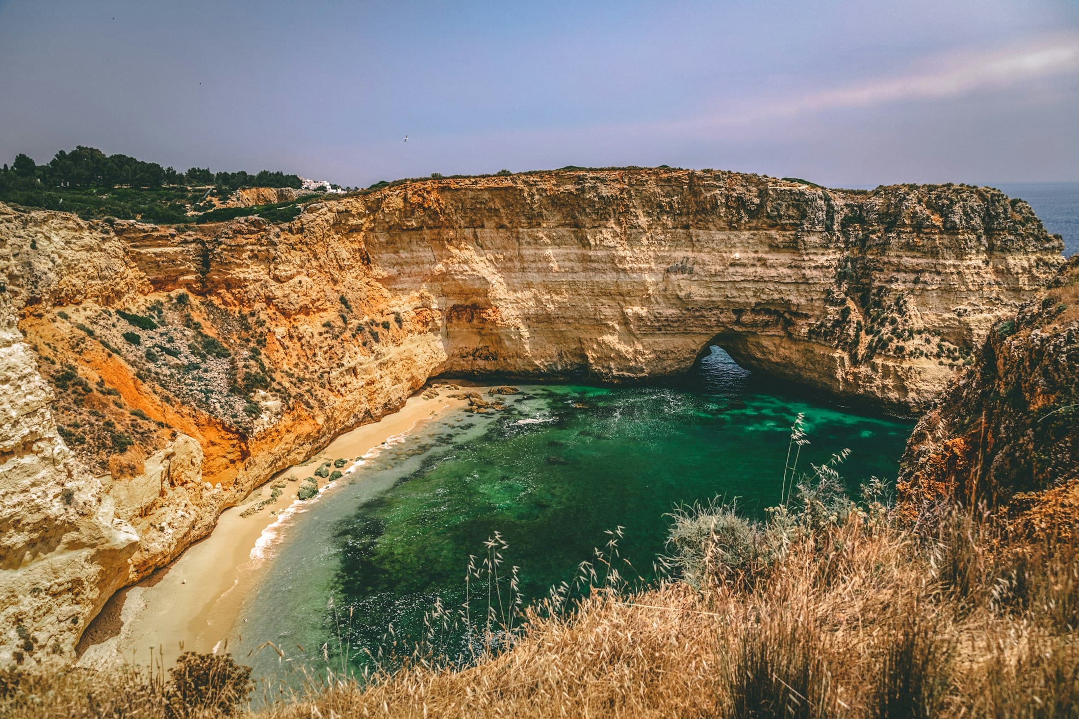 A hidden cove near Carvoeiro: sandstone cliffs, an arch open to the sea and green water below the dry grass of the cliff top