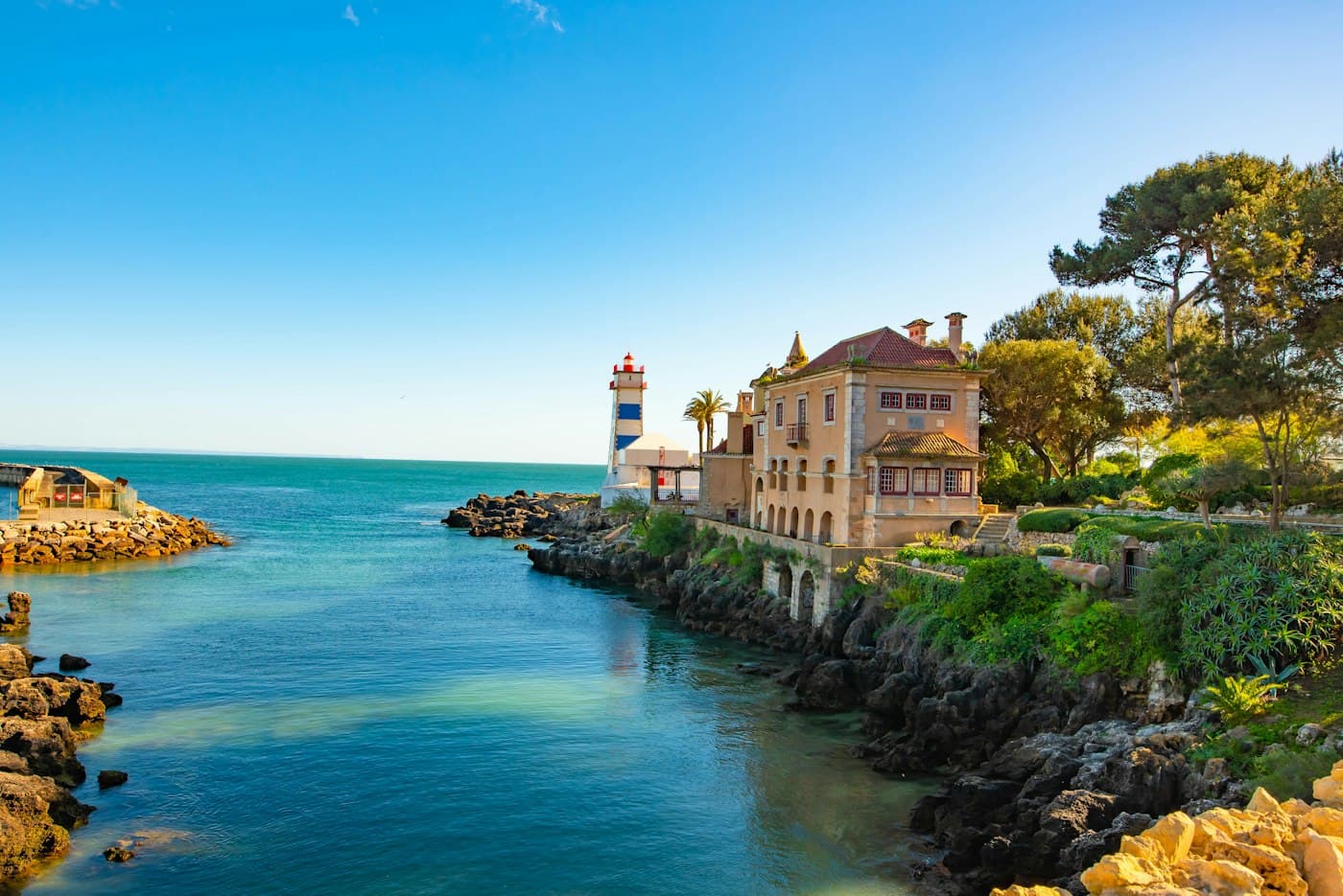 O farol de Santa Marta, às riscas azuis e brancas, e uma casa antiga entre pinheiros sobre uma enseada turquesa em Cascais