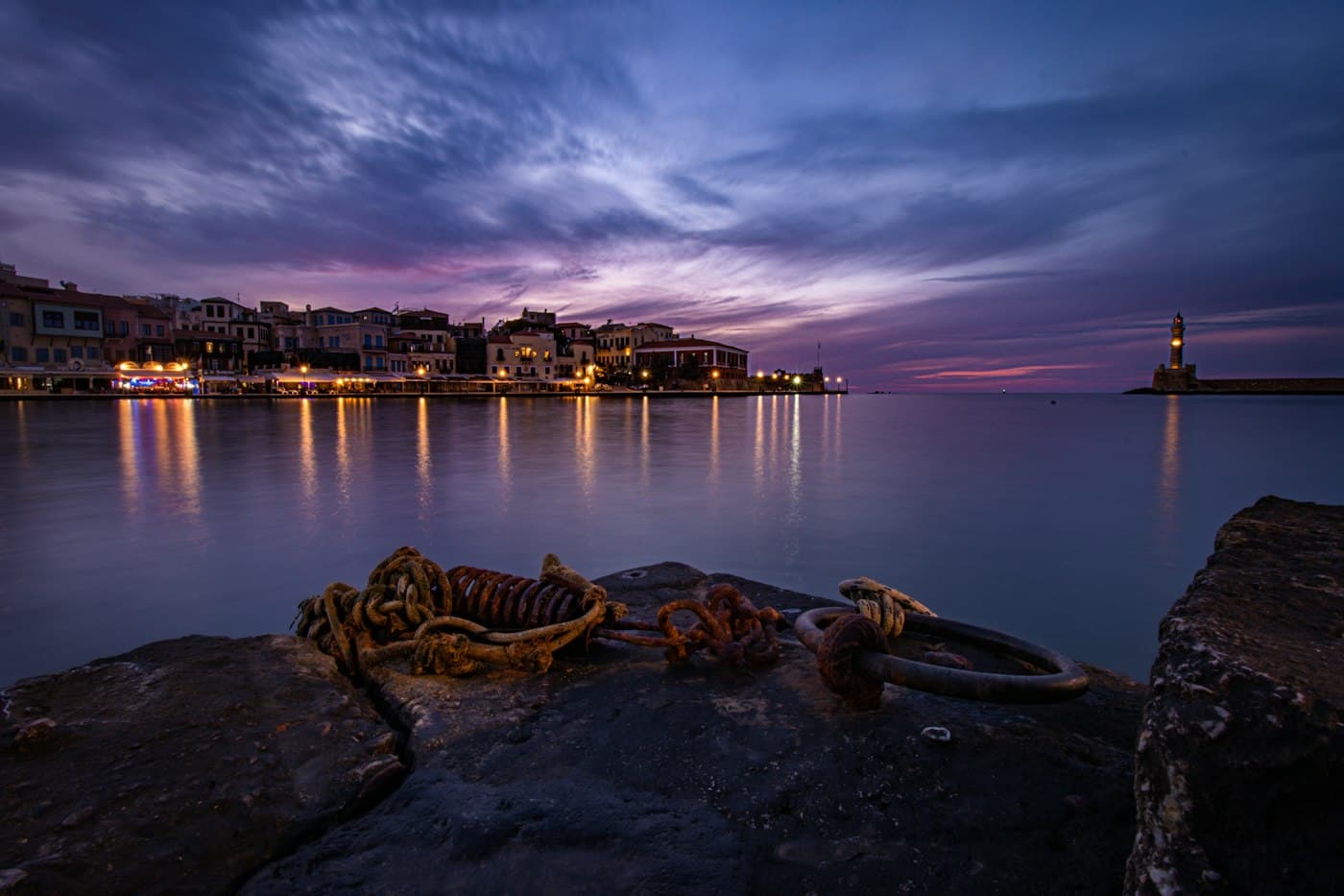 Chania’s harbour at blue hour from the old quay, the waterfront lit and the lighthouse glowing at the far end, mooring rings in the foreground