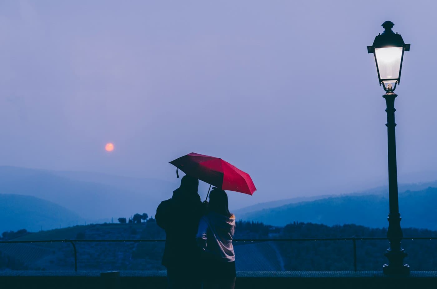 A couple under a red umbrella at a viewpoint in Radda in Chianti at dusk, looking out over misty hills