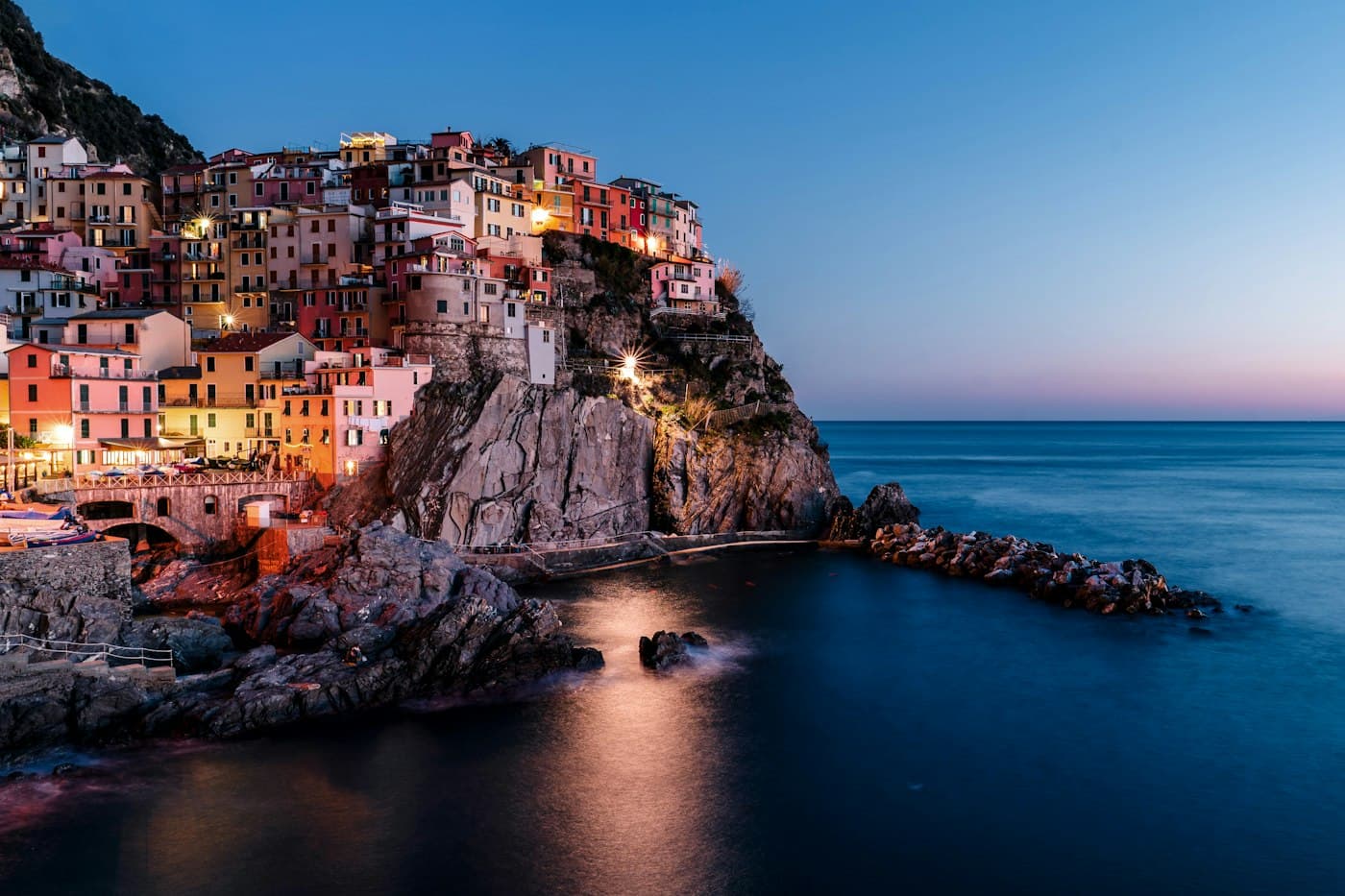 Manarola nell’ora blu, le case rosa, gialle e rosse una sopra l’altra su uno scoglio scuro sopra il mare calmo, i primi lampioni accesi lungo il camminamento