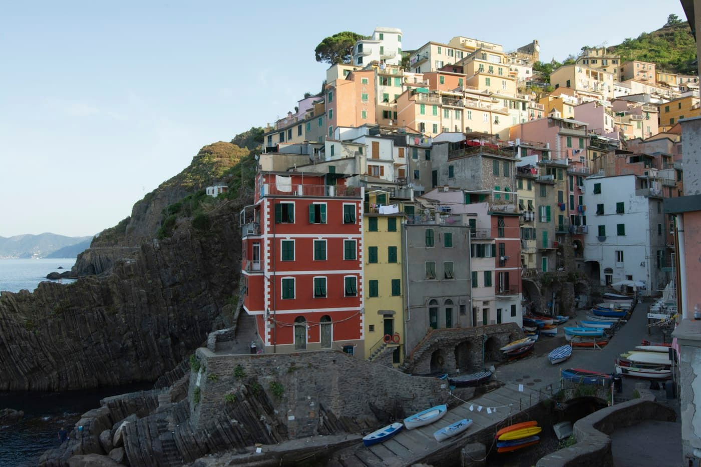 Riomaggiore nel primo sole: case alte, rosse, gialle e grigie che salgono sulla collina sopra lo scivolo, piccole barche tirate in secco sulle pietre