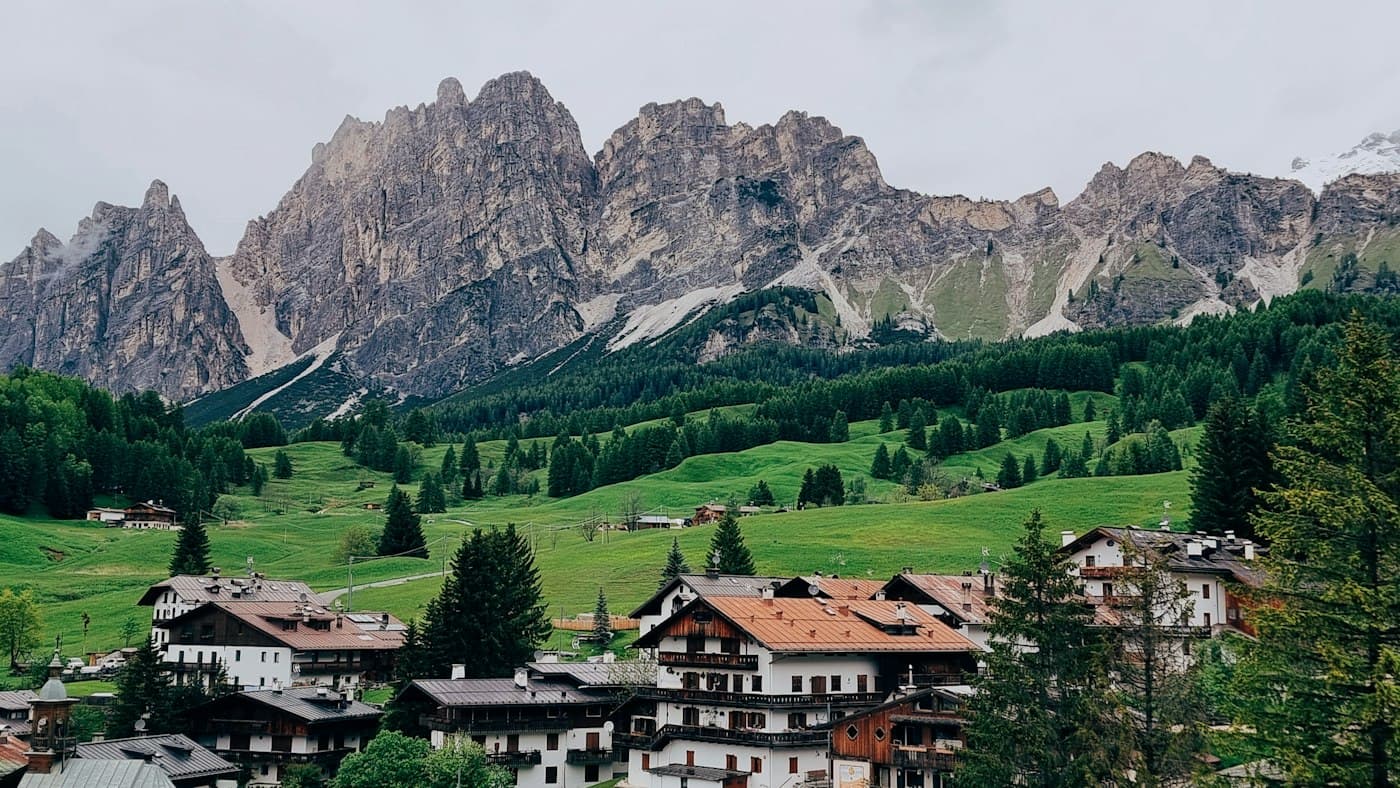 Chalet con muri bianchi, balconi di legno e tetti rossi ai margini di Cortina d’Ampezzo, prati verdi e boschi di abeti che salgono verso una cresta grigia e frastagliata