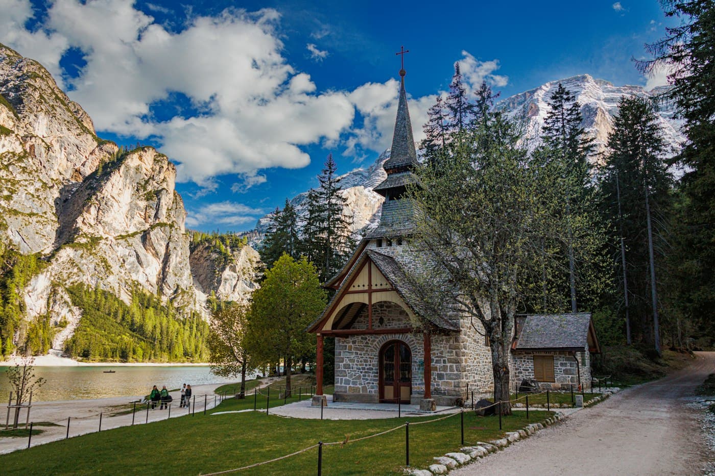La piccola cappella di pietra con la guglia di scandole sulla riva del Lago di Braies, tra larici e abeti, con una cima innevata alle spalle