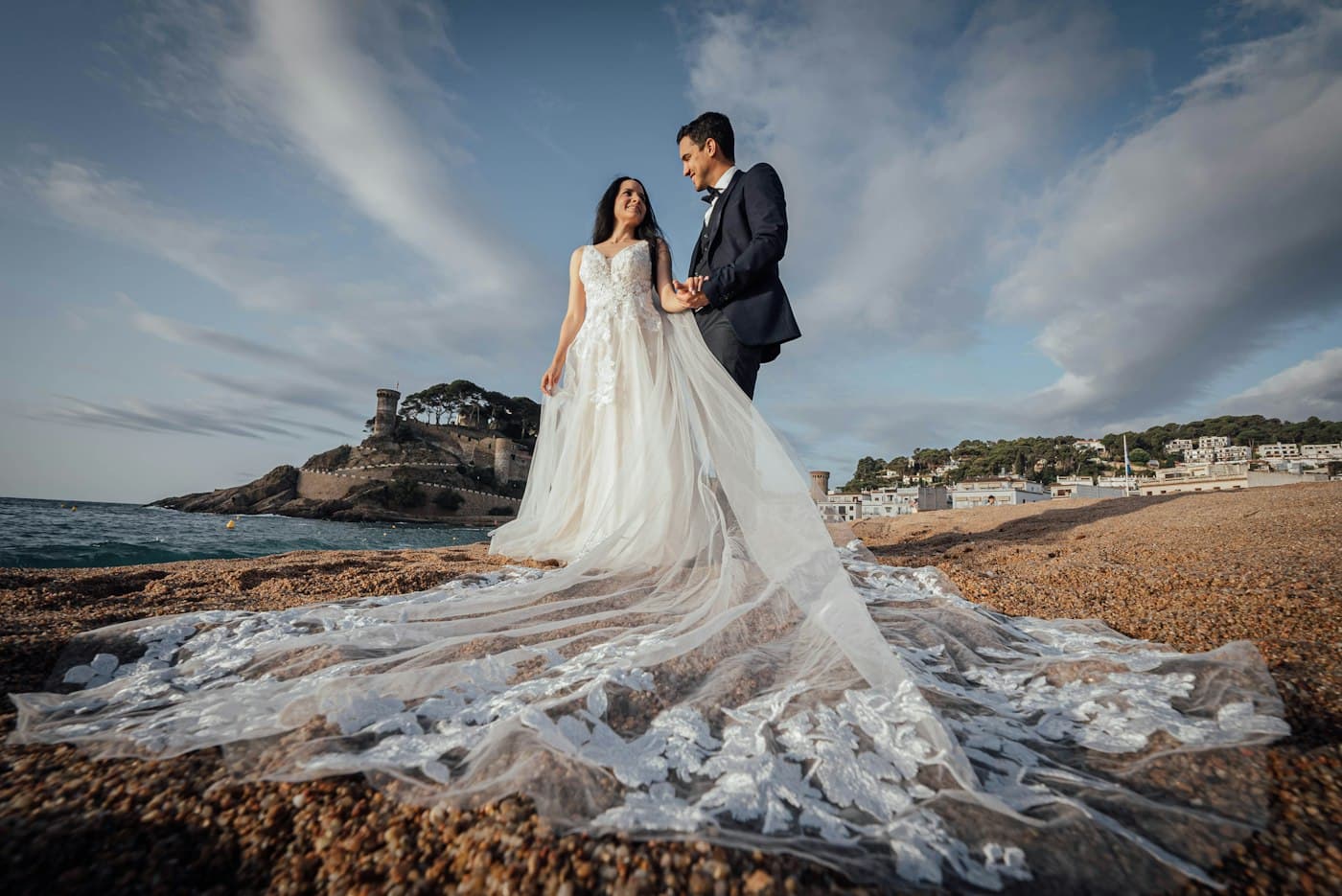 A bride and groom on the beach at Tossa de Mar, the long lace train of her dress spread over the sand, the walled town on its headland behind