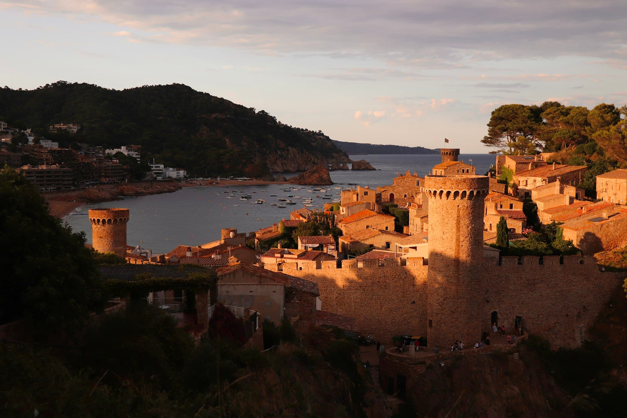 Las murallas y torres medievales de Tossa de Mar al atardecer, con la bahía y sus barcas detrás