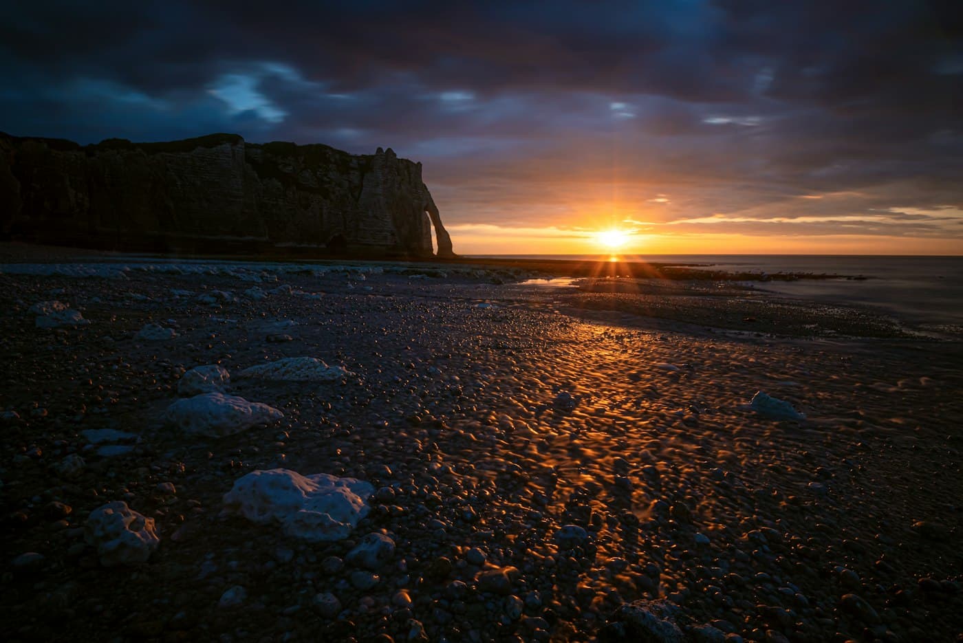 The sun setting over the sea beside the dark outline of the Porte d’Aval, the pebbles of Étretat’s beach lit orange at low tide