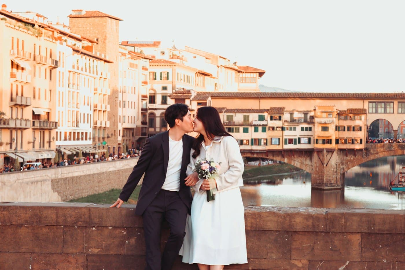Una sposa con il bouquet e lo sposo si baciano su un parapetto di pietra lungo l’Arno, alle loro spalle il Ponte Vecchio