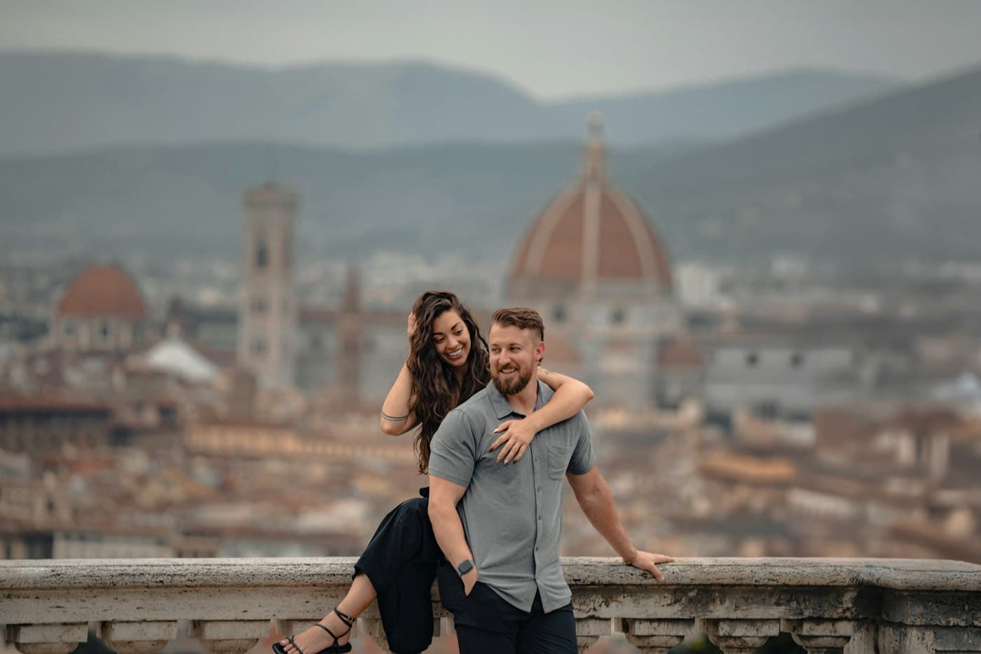 Una coppia sorride appoggiata a una balaustra di pietra sopra Firenze, alle spalle la cupola del Duomo e il campanile sfocati