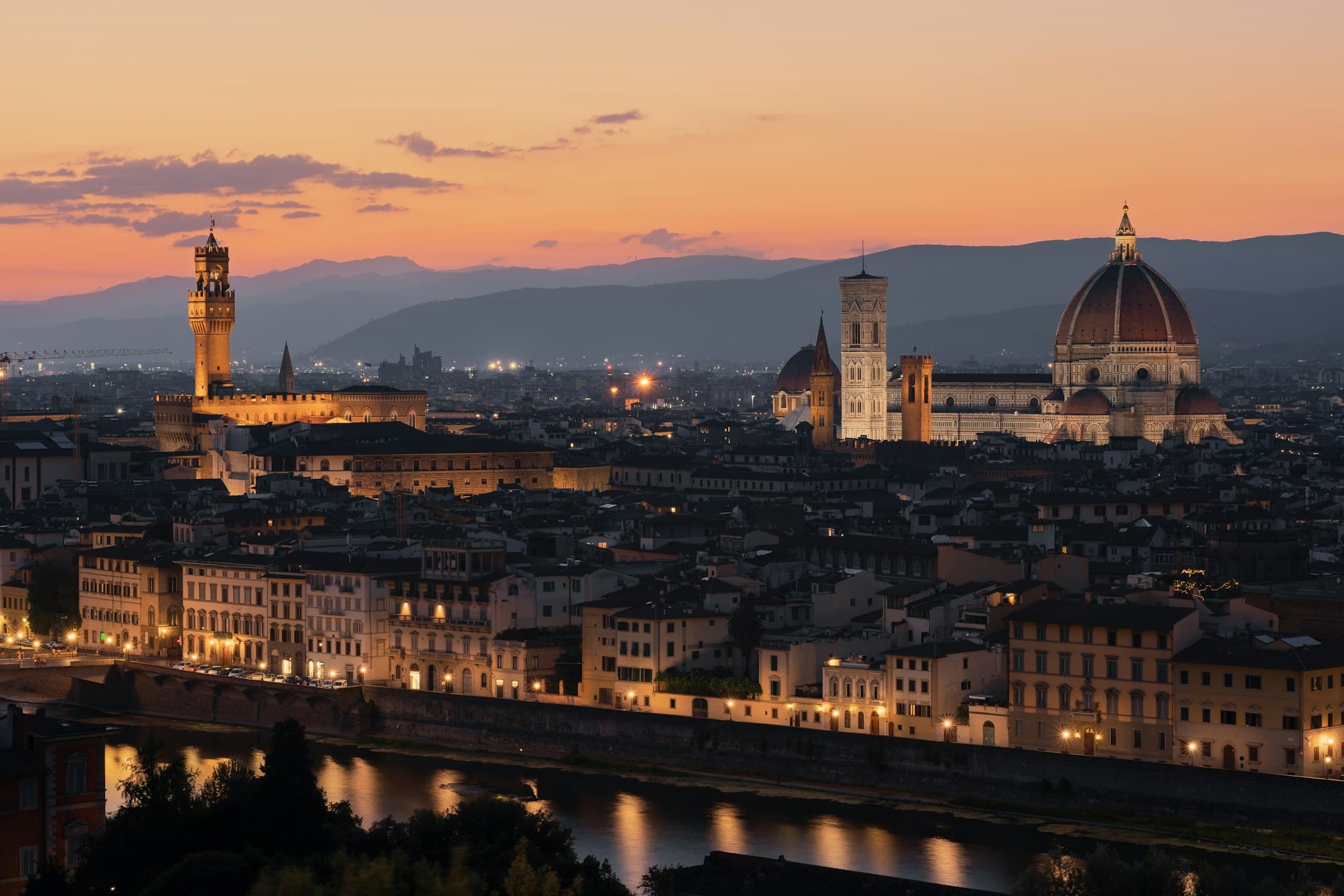 Florence at dusk seen from above the Arno: the lit tower of Palazzo Vecchio and the cathedral dome over the rooftops