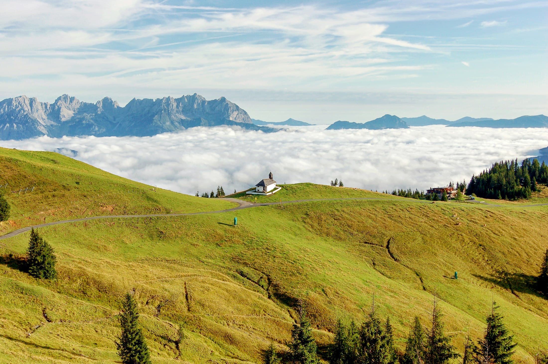 A small white chapel on the green slopes of the Hahnenkamm above a sea of fog, the jagged Wilder Kaiser rising behind