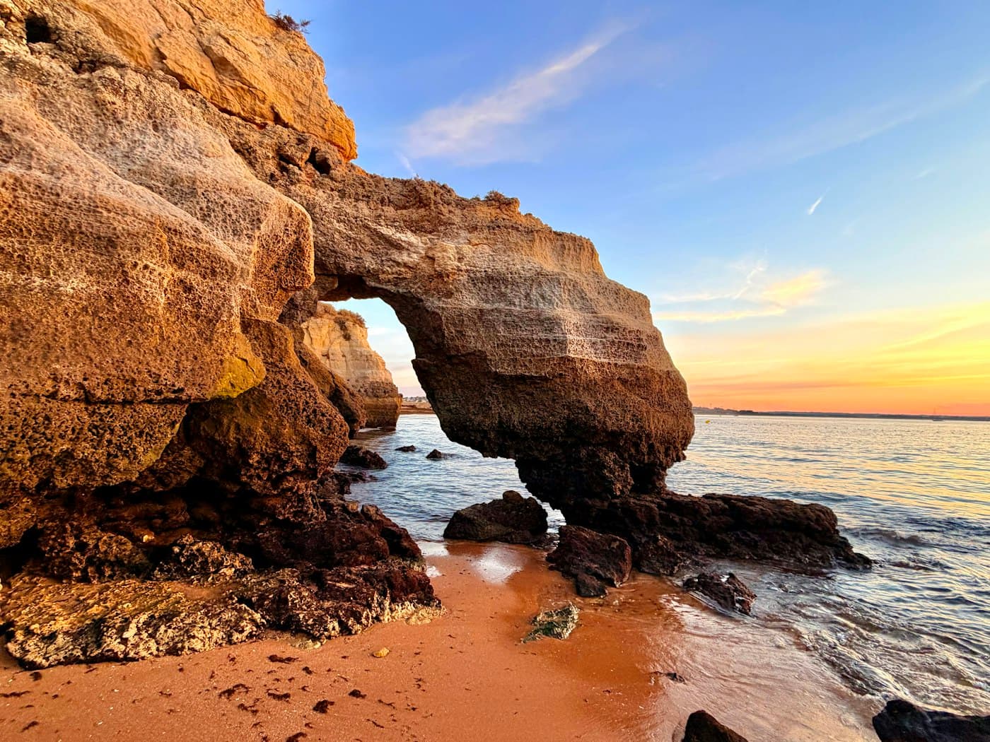 A sandstone arch on a small beach in Lagos at sunset, the sky turning orange over the bay