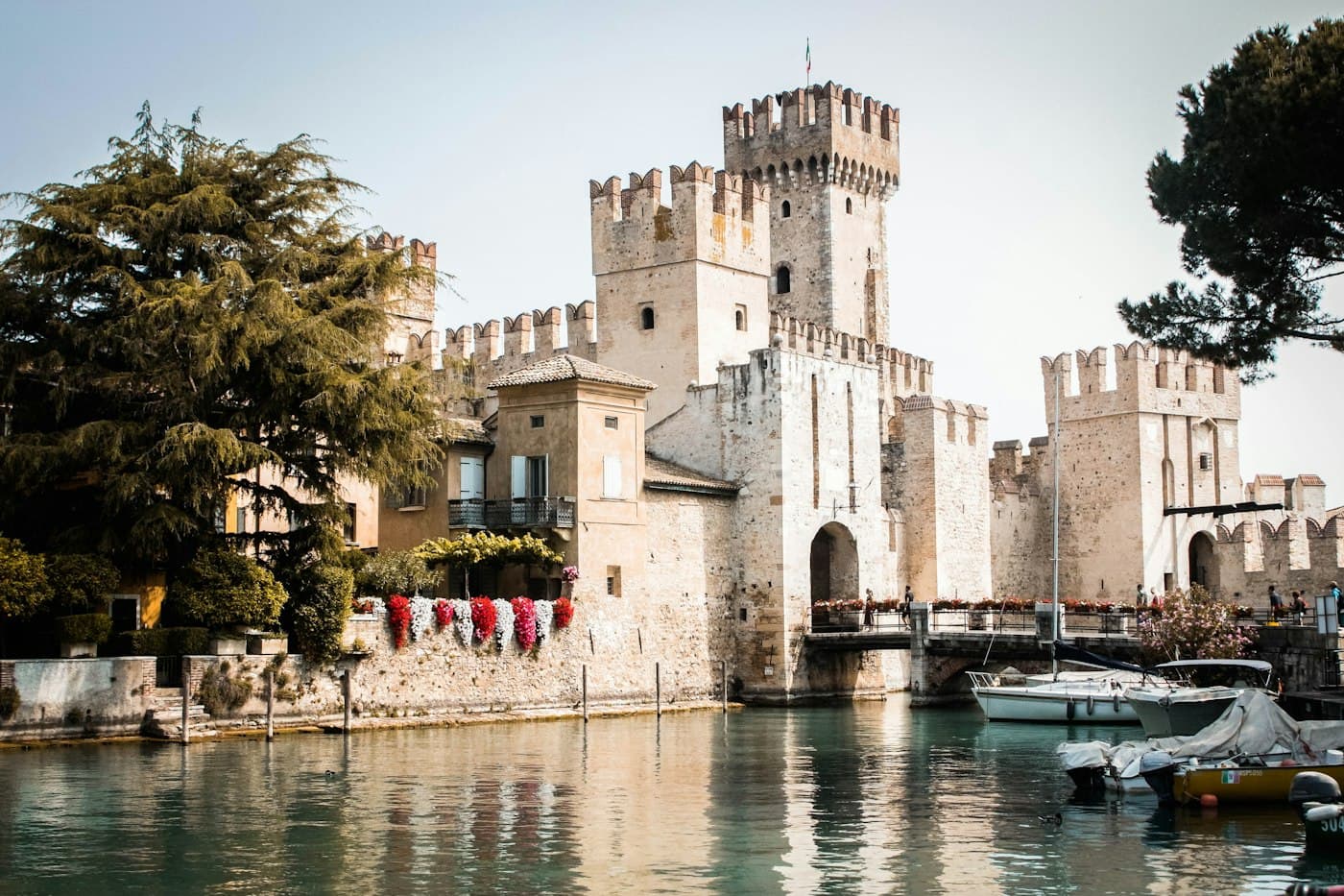 Il castello scaligero di Sirmione che sorge dall’acqua, con fiori lungo il muro e barche ormeggiate accanto al ponte