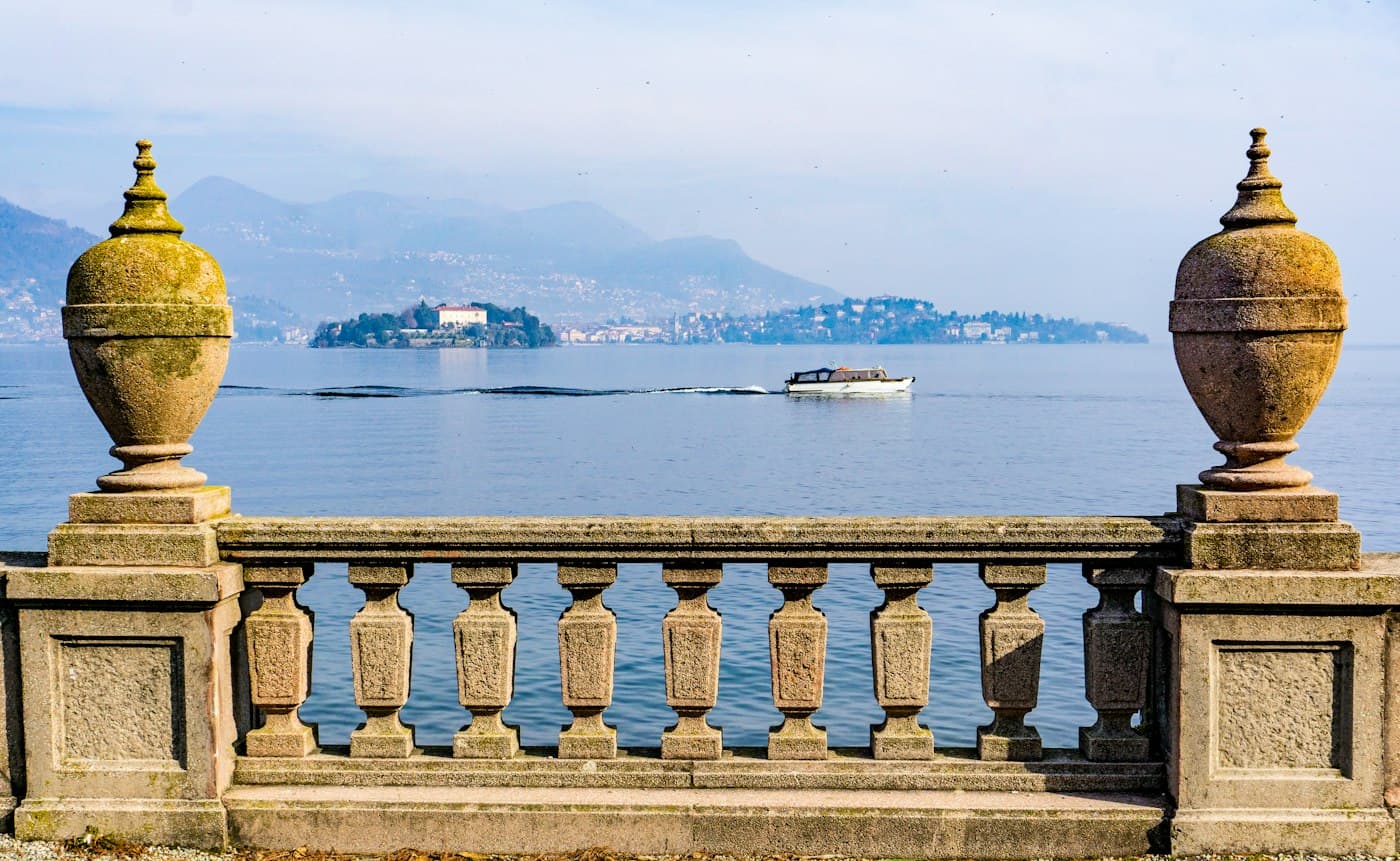 Due vasi di pietra su una balaustra di Stresa che incorniciano il lago velato, un motoscafo che attraversa verso l’Isola Madre e le colline dell’altra sponda