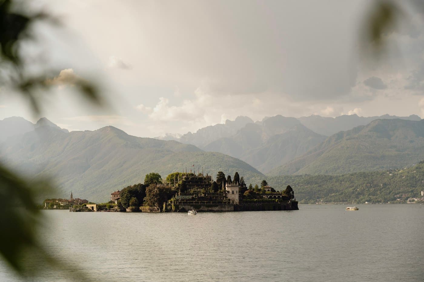 L’Isola Bella vista dalla riva tra le foglie, il palazzo e i giardini a terrazze che salgono dall’acqua grigia, le montagne nella foschia alle spalle