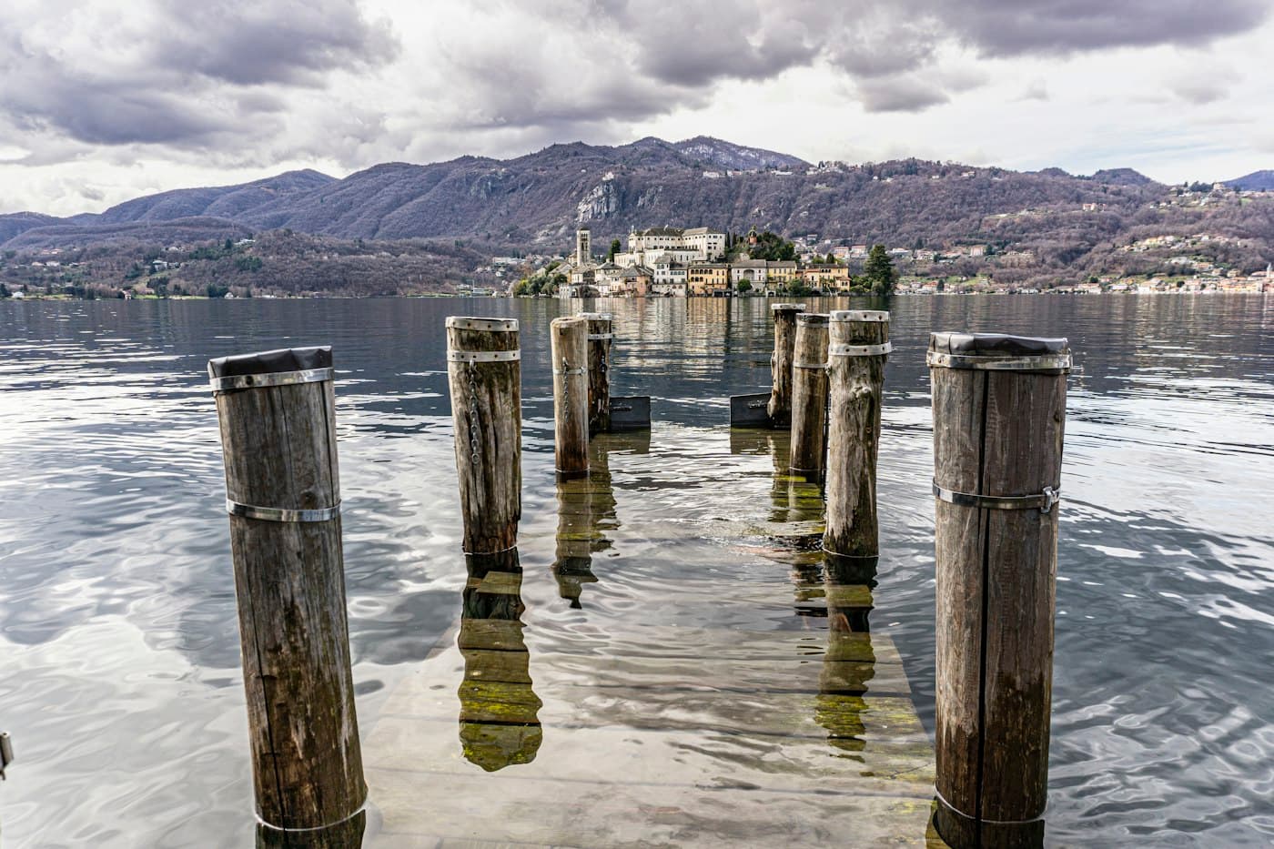 Old wooden mooring posts standing in the lake, the island of San Giulio and the mountains behind under a cloudy sky