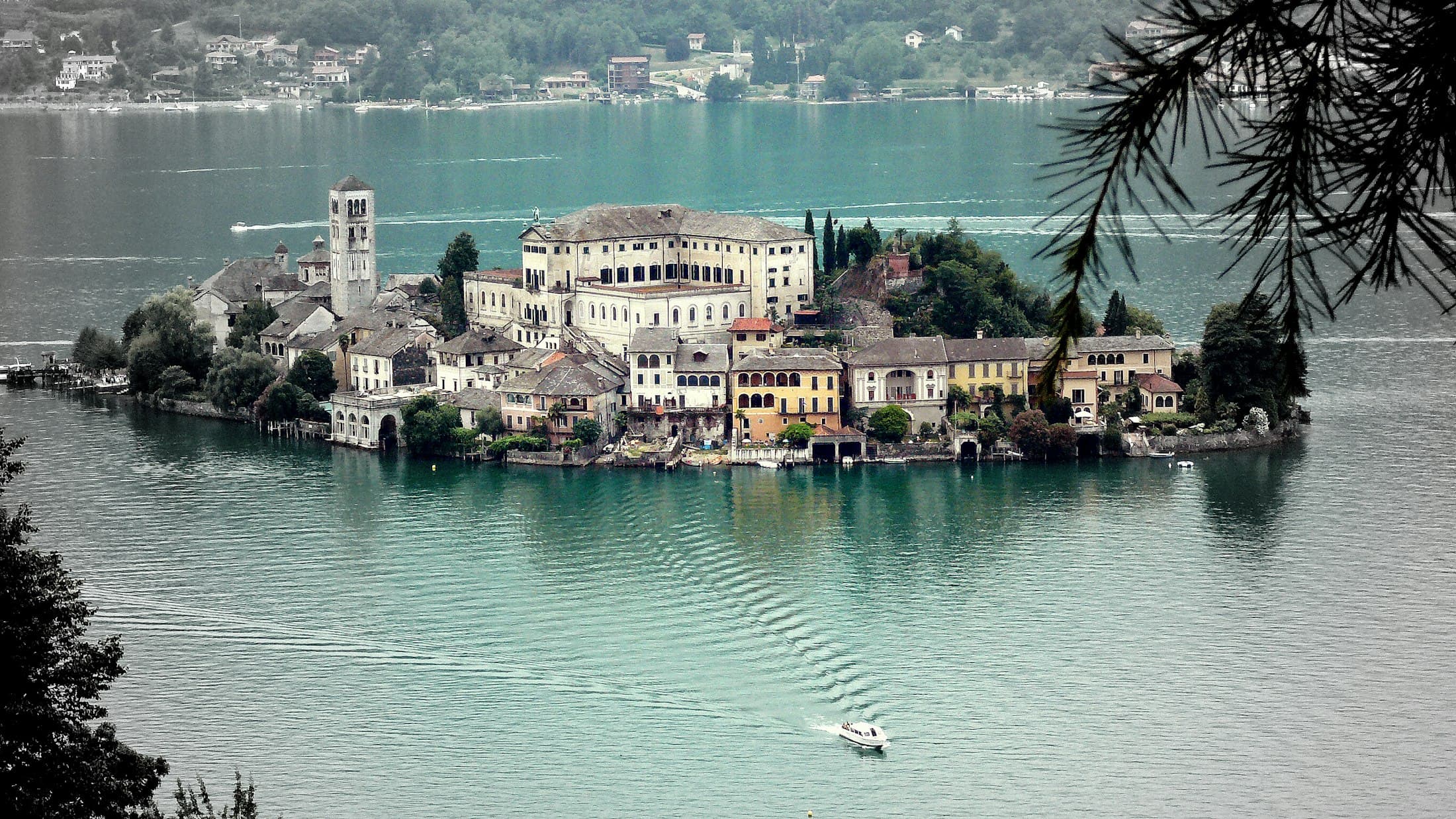 L’isola di San Giulio vista dall’alto, il campanile e gli edifici chiari circondati dall’acqua verde, con una barca che le si avvicina