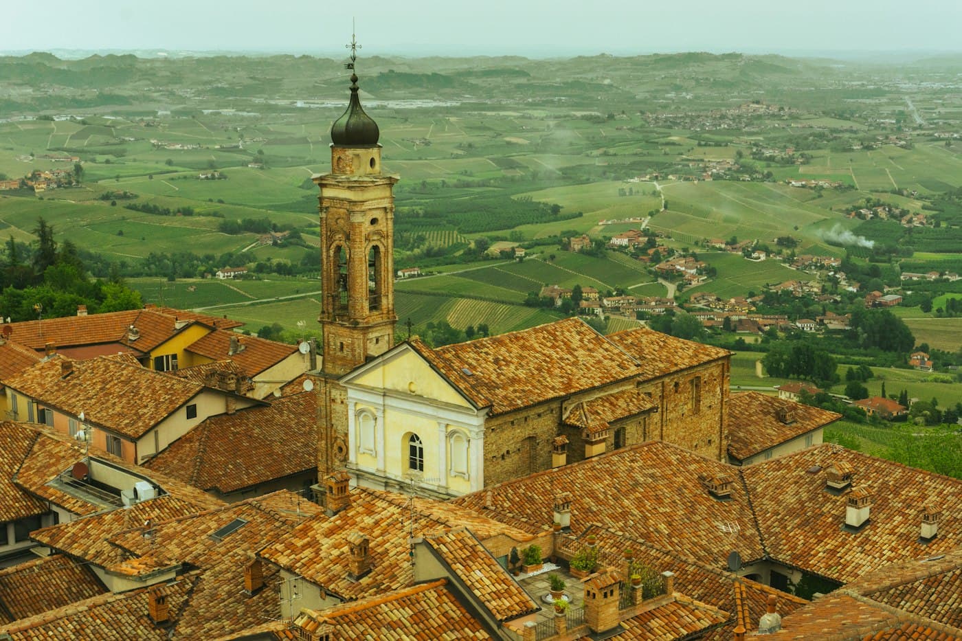 Un campanile sopra i tetti di coppi di La Morra, con i vigneti e i paesi delle Langhe che si stendono più in basso