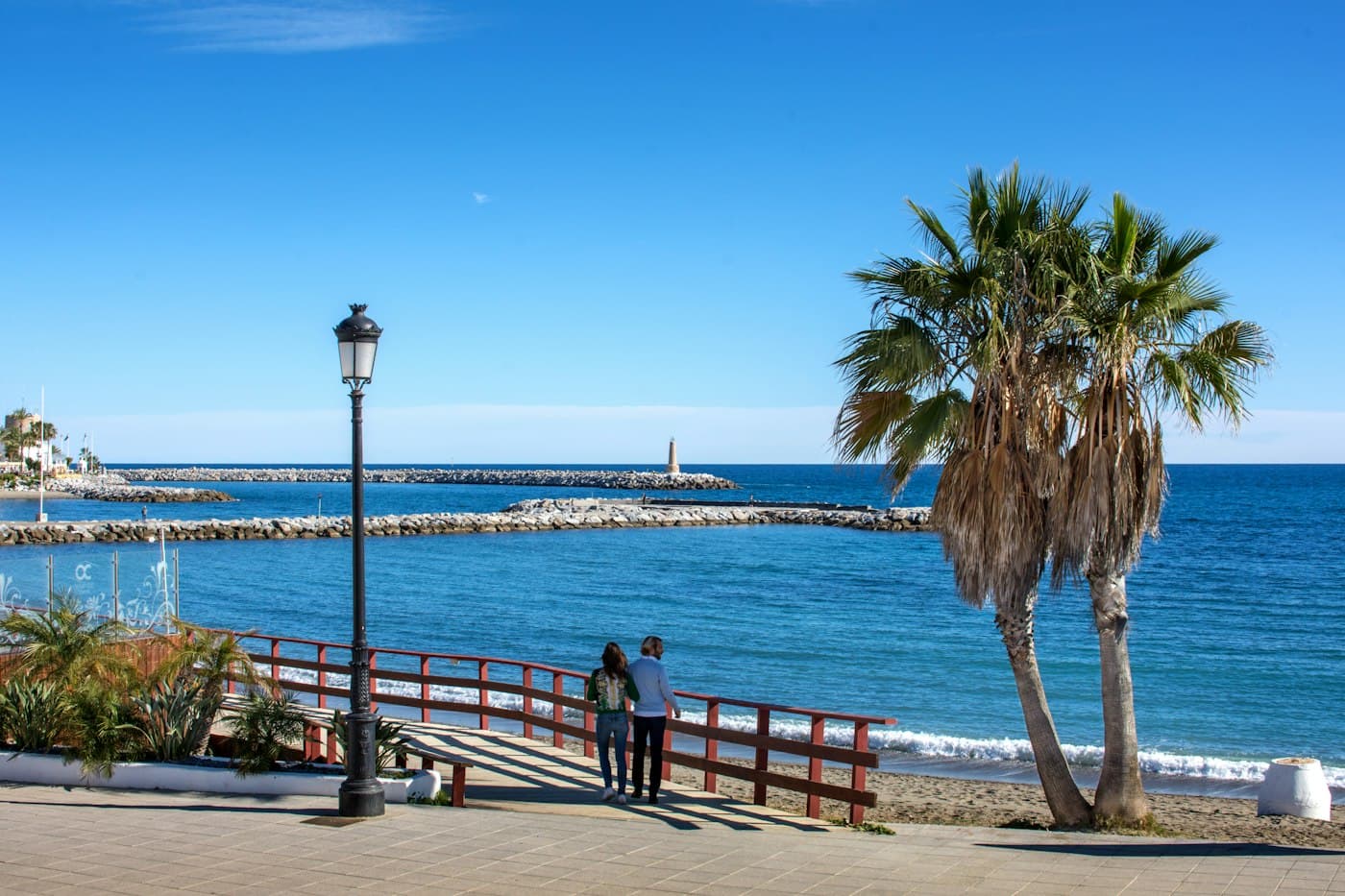 A couple at the railing of the seafront promenade in Puerto Banús, palm trees and the harbour wall behind