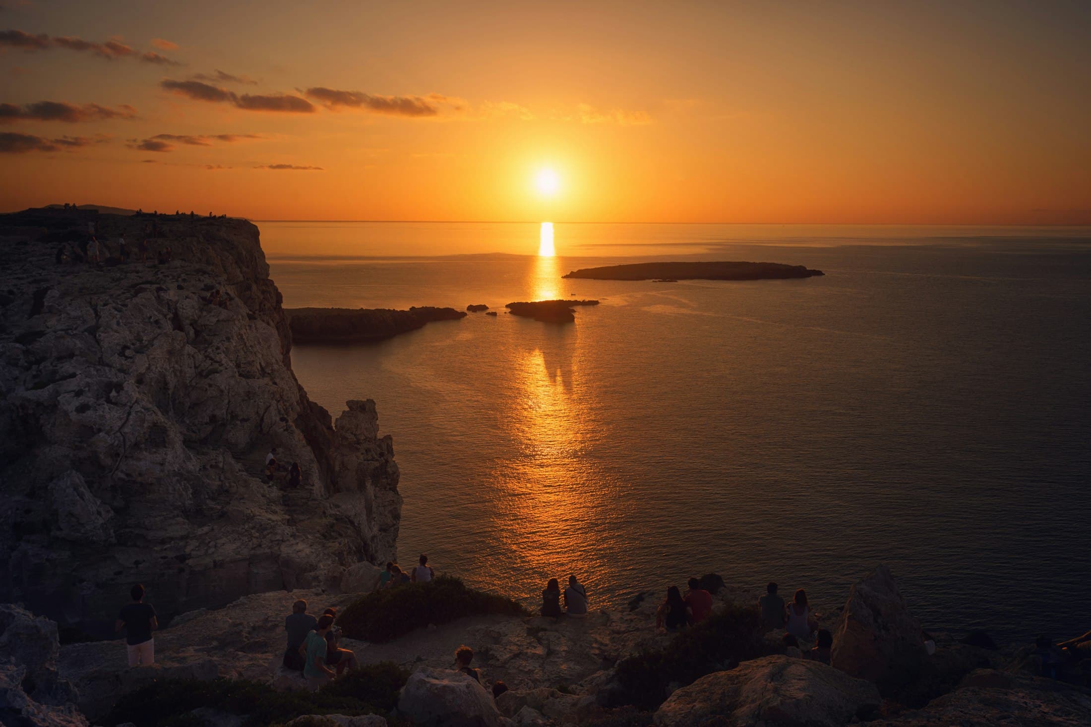 Gente en los acantilados junto al faro de Cavalleria, en el norte de Menorca, viendo ponerse el sol sobre el mar y sus islotes