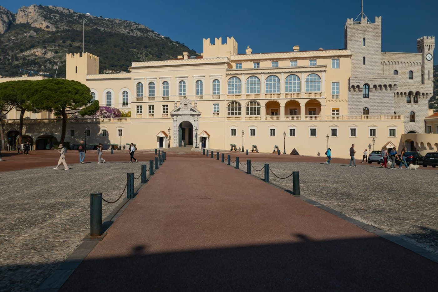 The Prince’s Palace of Monaco across its square in bright sun, a walkway between chained bollards leading to the gate, the mountain behind