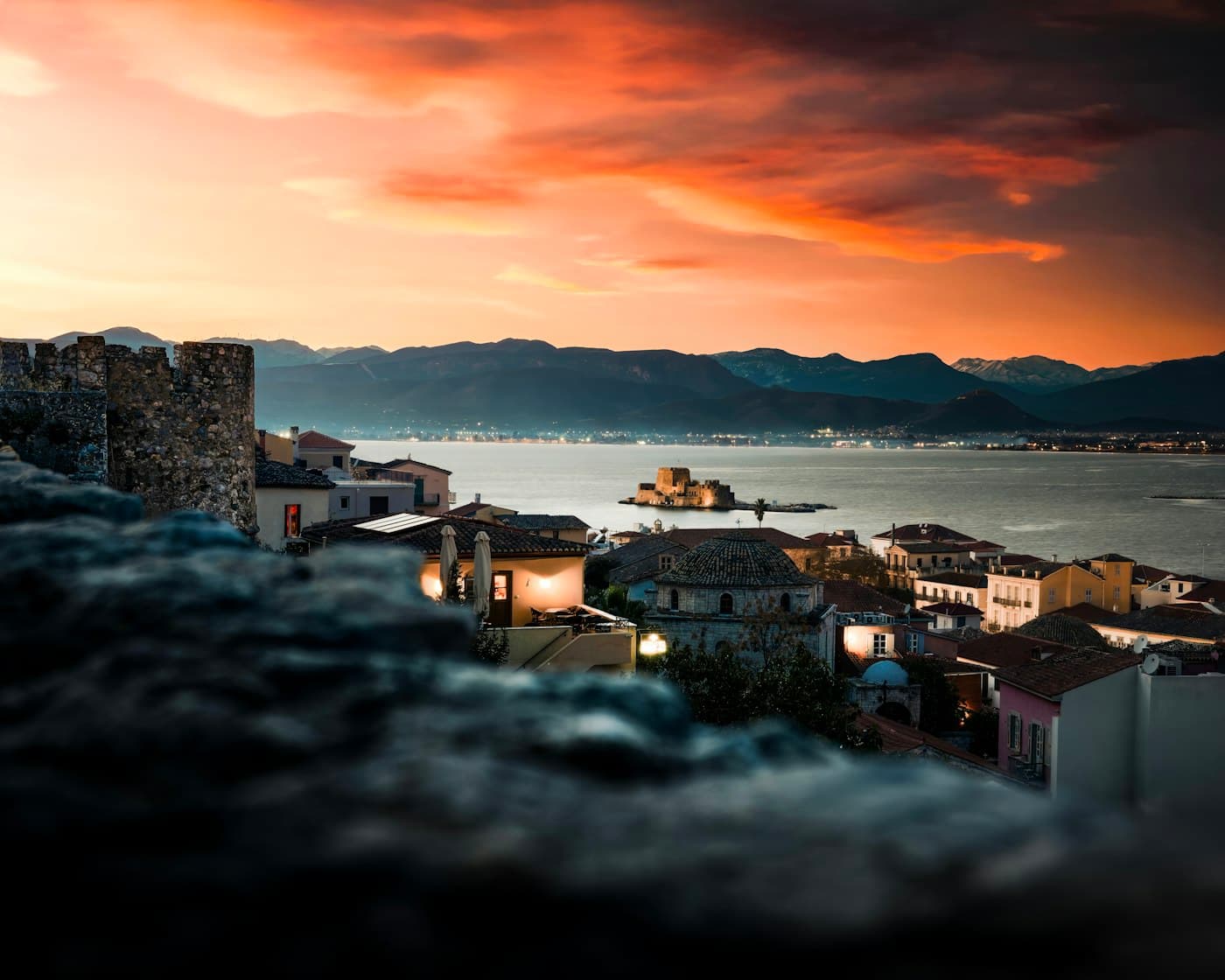 Sunset over Nafplio from the old fortress walls: lit houses, the Bourtzi in the bay and the mountains across the Argolic Gulf