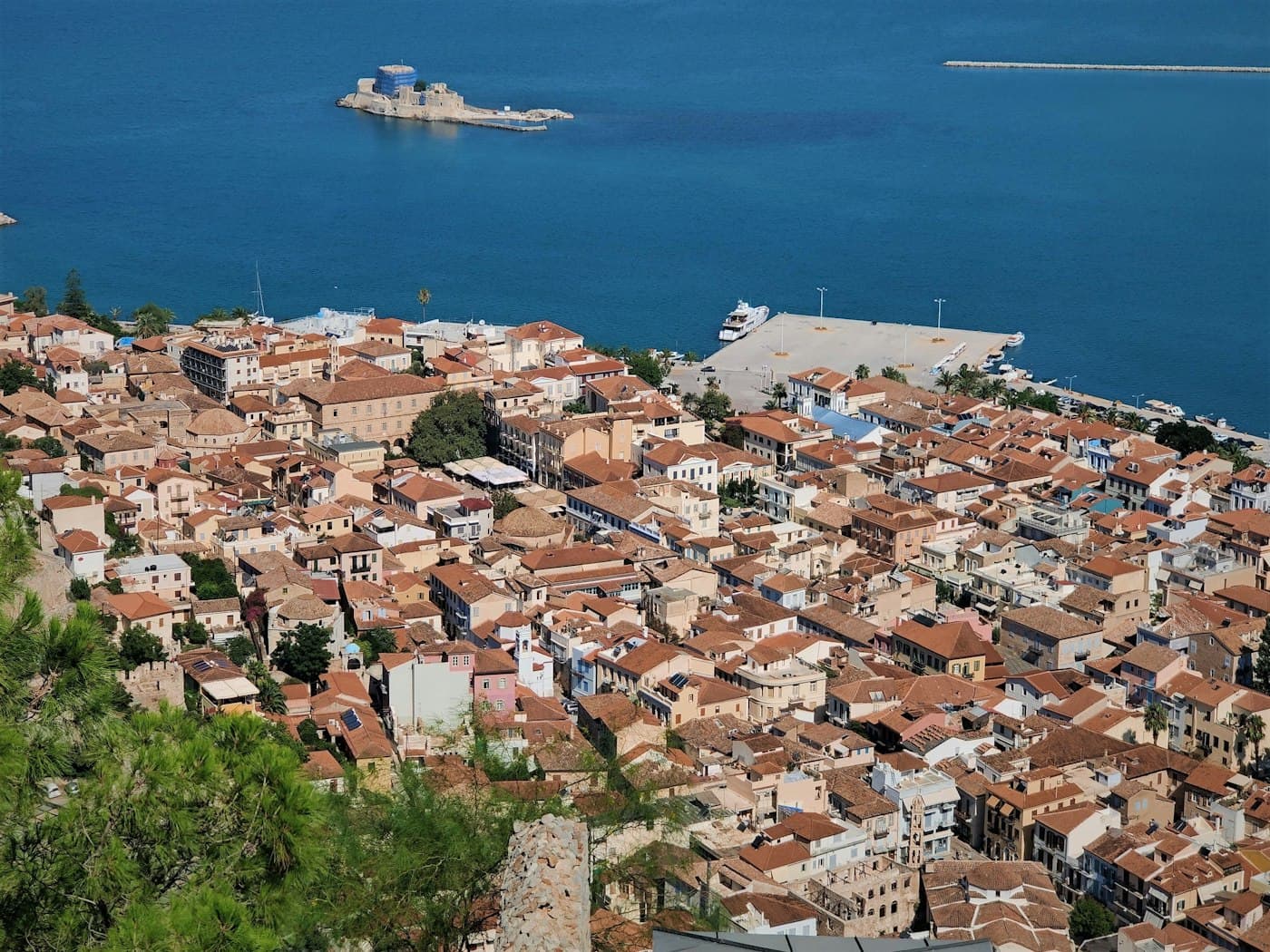 Nafplio’s old town from the Palamidi: tiled roofs down to the harbour, the Bourtzi on its islet in the bay, its tower wrapped for restoration