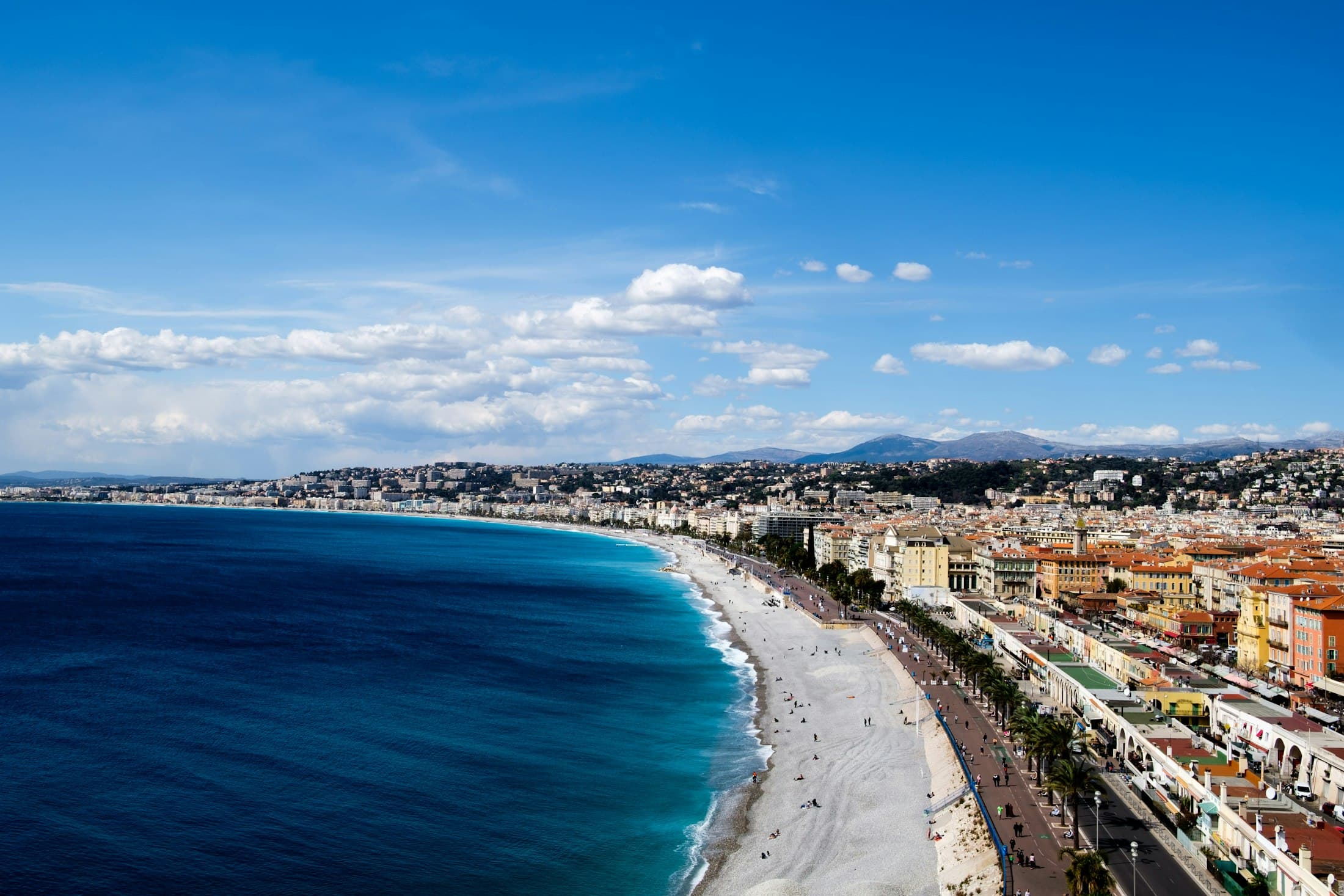 The Baie des Anges from Castle Hill: the pebble beach and the palm-lined Promenade des Anglais curving away along the city, the hills behind