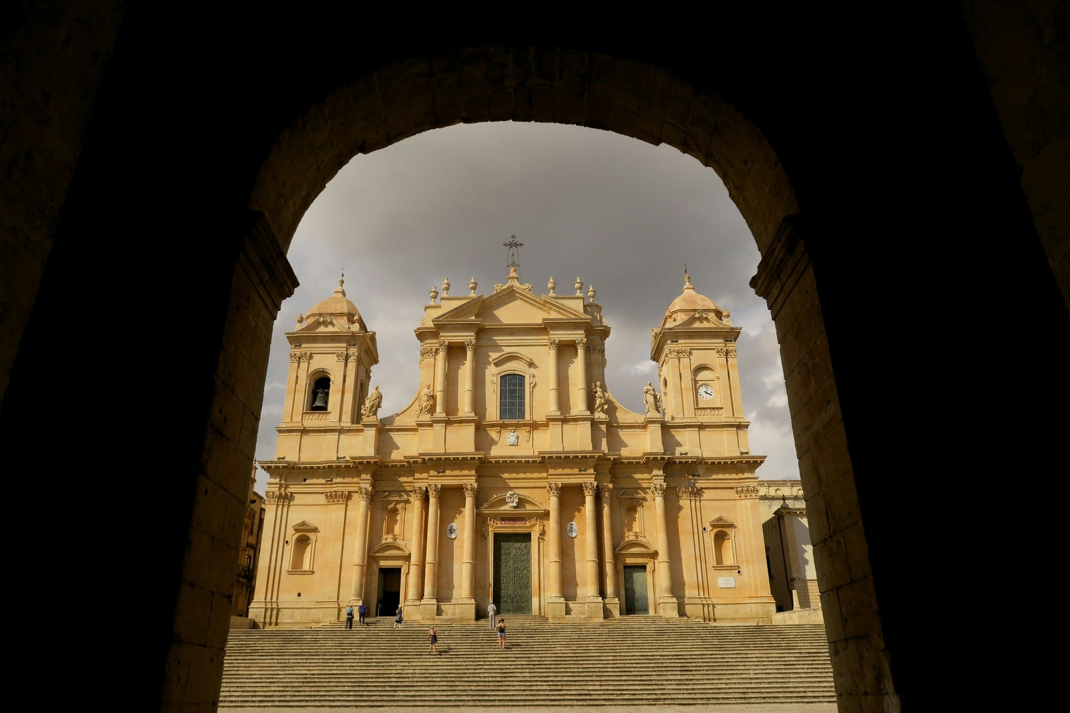 La cattedrale barocca di Noto e la sua ampia scalinata, incorniciate da un arco di pietra in ombra