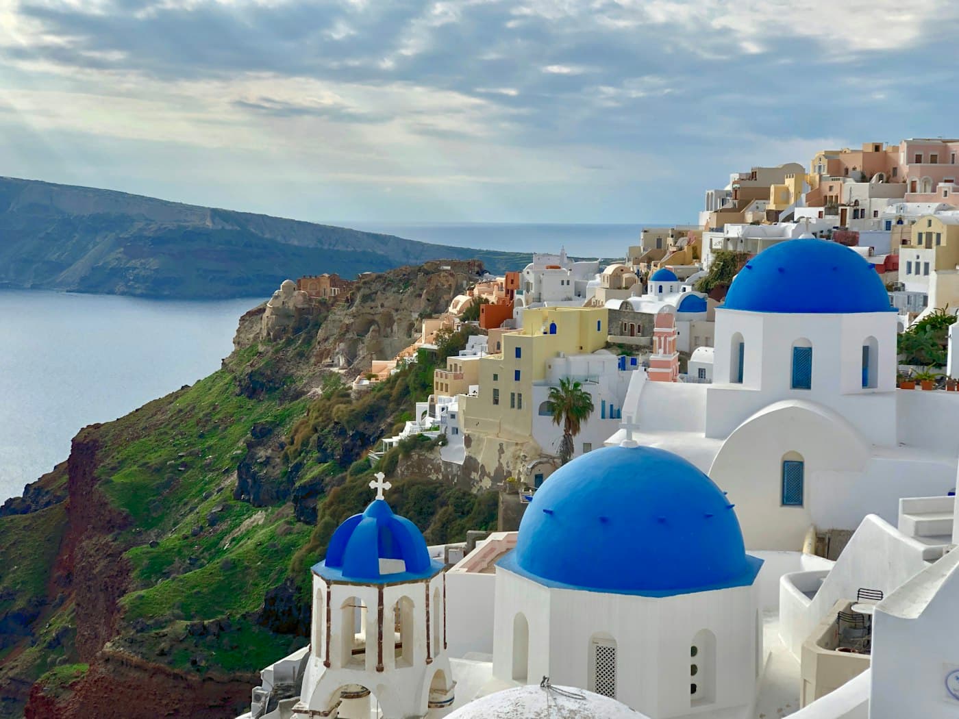 Two blue-domed churches and a bell tower in Oia, the caldera’s green and red slopes falling to the sea beside them